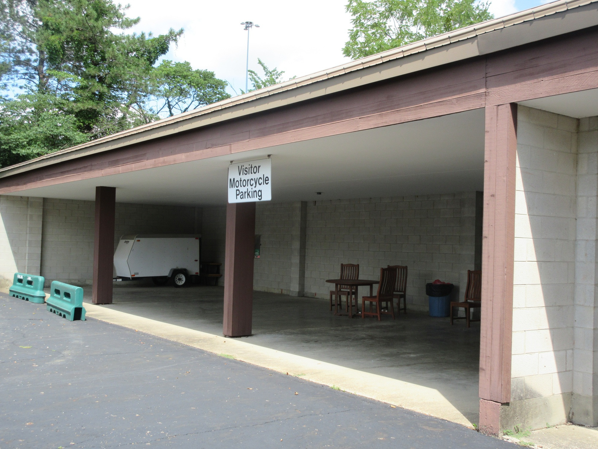 Covered parking area with a sign saying "motorcycle parking" that is empty except for a small trailer and some chairs and a table.