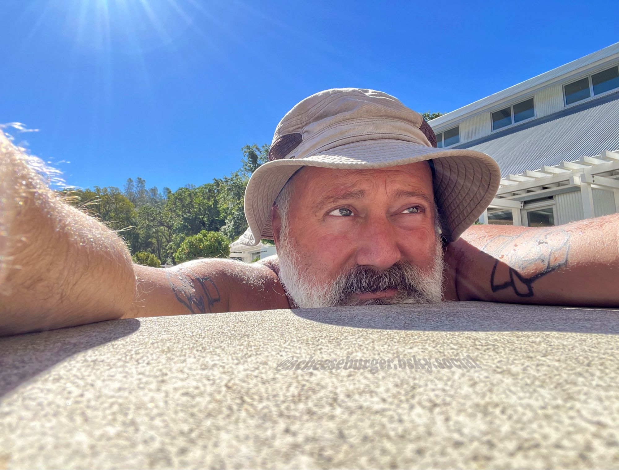 grey bearded guy with a full brim hat hanging on the edge of a pool on a sunny day.