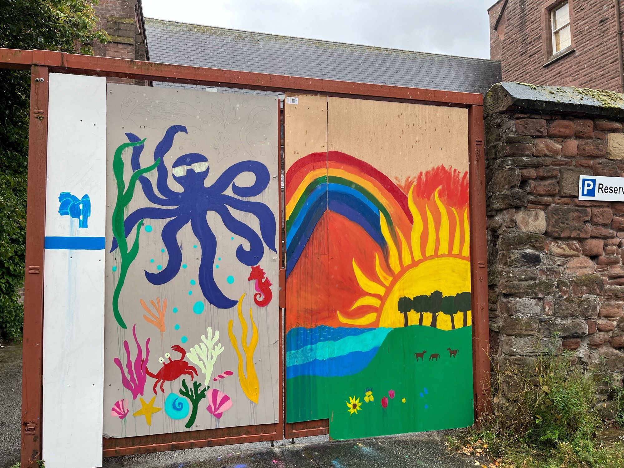 A boarded up gate has been painted with murals in bright colours. A purple octopus & a rainbow sunset.