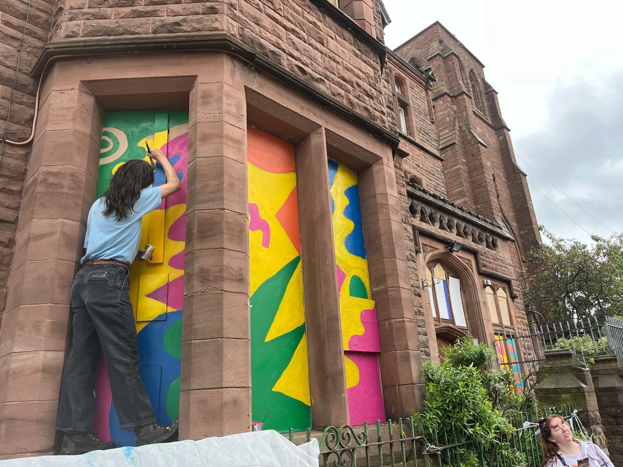 Artist standing on the window ledge of a sandstone Victorian building. Painting the boards over windows in bright abstract patterns.