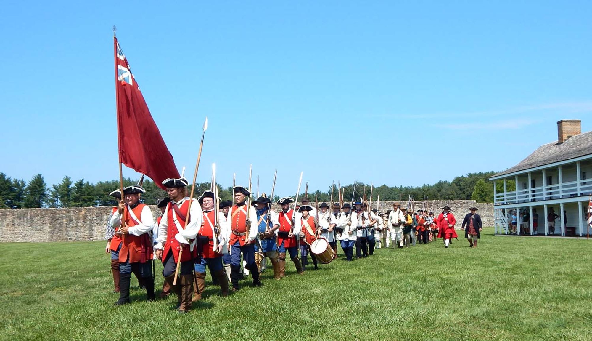 A column of assorted colonial provincial troops march from the Fort Frederick barracks and across the parade ground to seek out French troops in the vicinity. They wear a variety of colonial American attire, notably black tricorn hats, with most carrying shouldered muskets. A flag bearer (carrying the red provincial flag with Union Jack in the corner) and an officer with a spontoon lead the column, with a drummer not far behind.