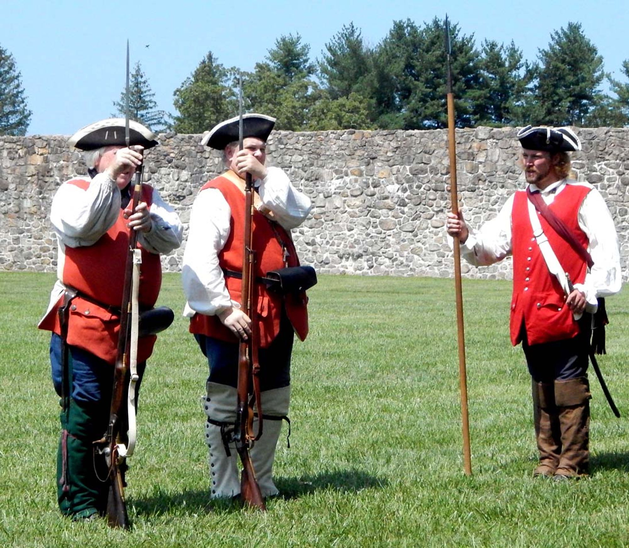 Two members of Mercer’s Provincial Company fix bayonets before starting guard duty at the fort barracks. They wear black tricorn hats, white shirts and a red waistcoat over breeches and gaiters, each affixing a bayonet to the end of their musket, braced on the ground. Their sergeant is similarly attired and holding a spontoon (a spear-like implement used more as an indication of rank and a means to direct commands).