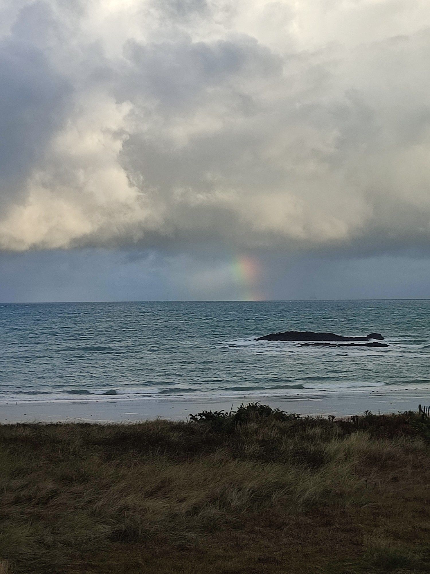 Pied d'arc en ciel visible au dessus de la mer et sous une couche de nuages gris et blancs