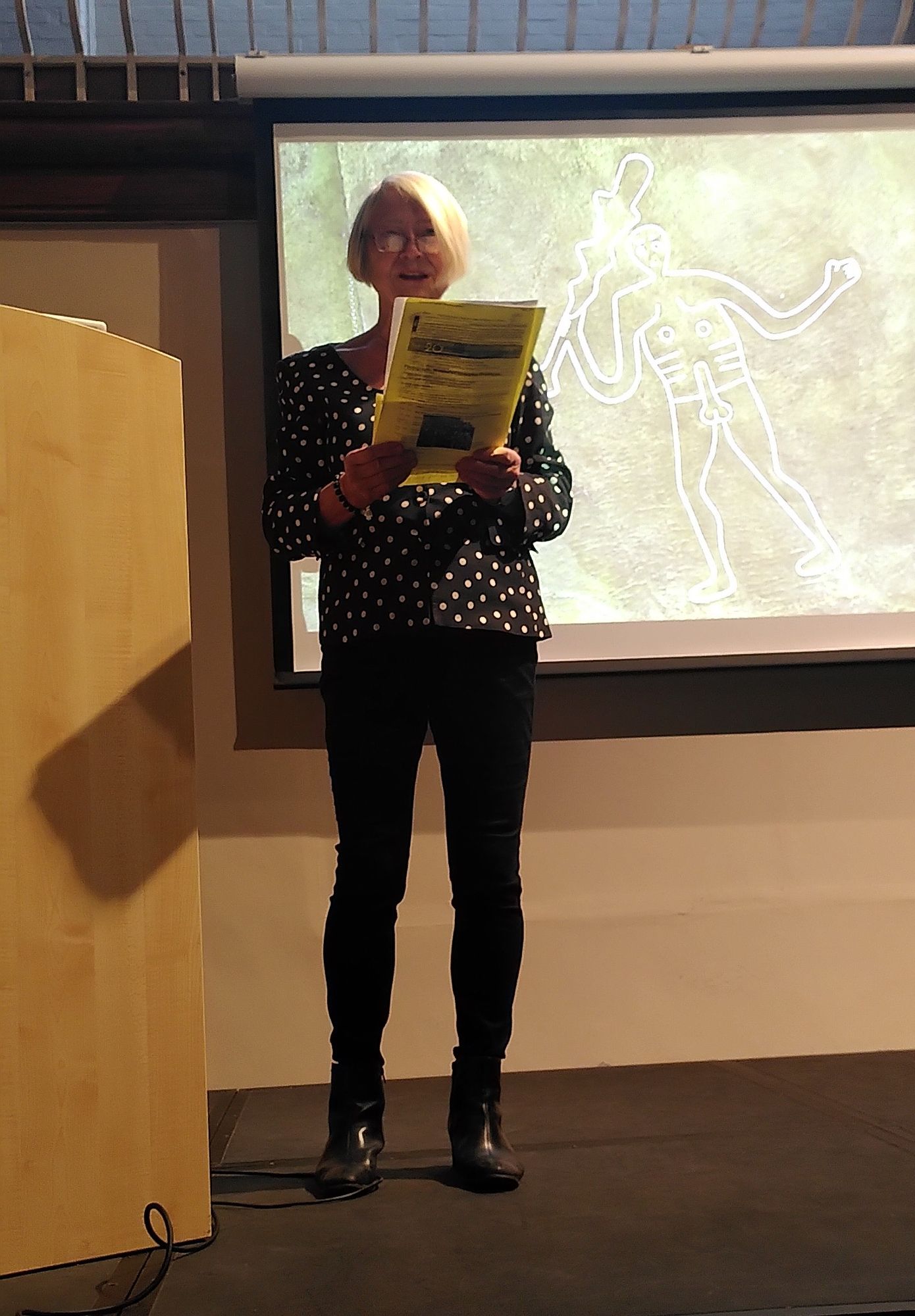 A woman with short bobbed blond hair and glasses, wearing a spotty top, black trousers and boots, stands on a stage speaking holding papers, in front of a projected photo of the Cerne Abbas Giant, a Saxon chalk hill figure