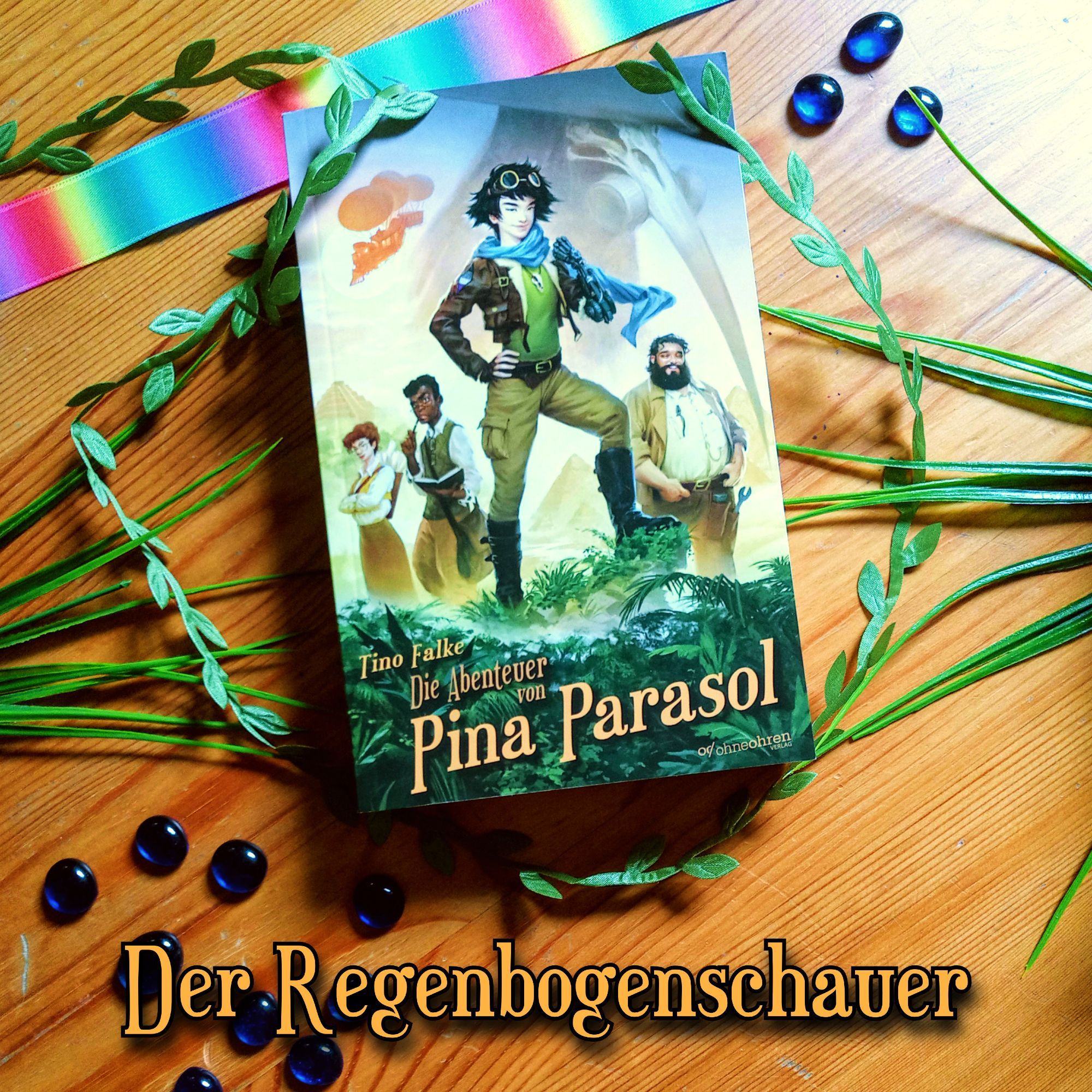 Foto vom Buch „Die Abenteuer von Pina Parasol“ vor braunem Holzhintergrund. Rund um das Buch ragen Plastikpflanzen ins Bild, am oberen Rand liegt ein regenbogenfarbenes Band, überall liegen kleine blaue Glassteine. Darunter steht in Braun: „Der Regenbogenschauer“.