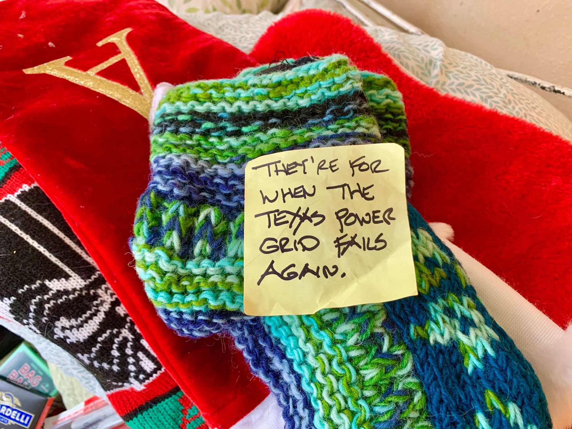 Extremely thick woolen slipper socks knitted in blue and green yarns, with a sticky note reading, “They’re for when the Texas power grid fails again.”