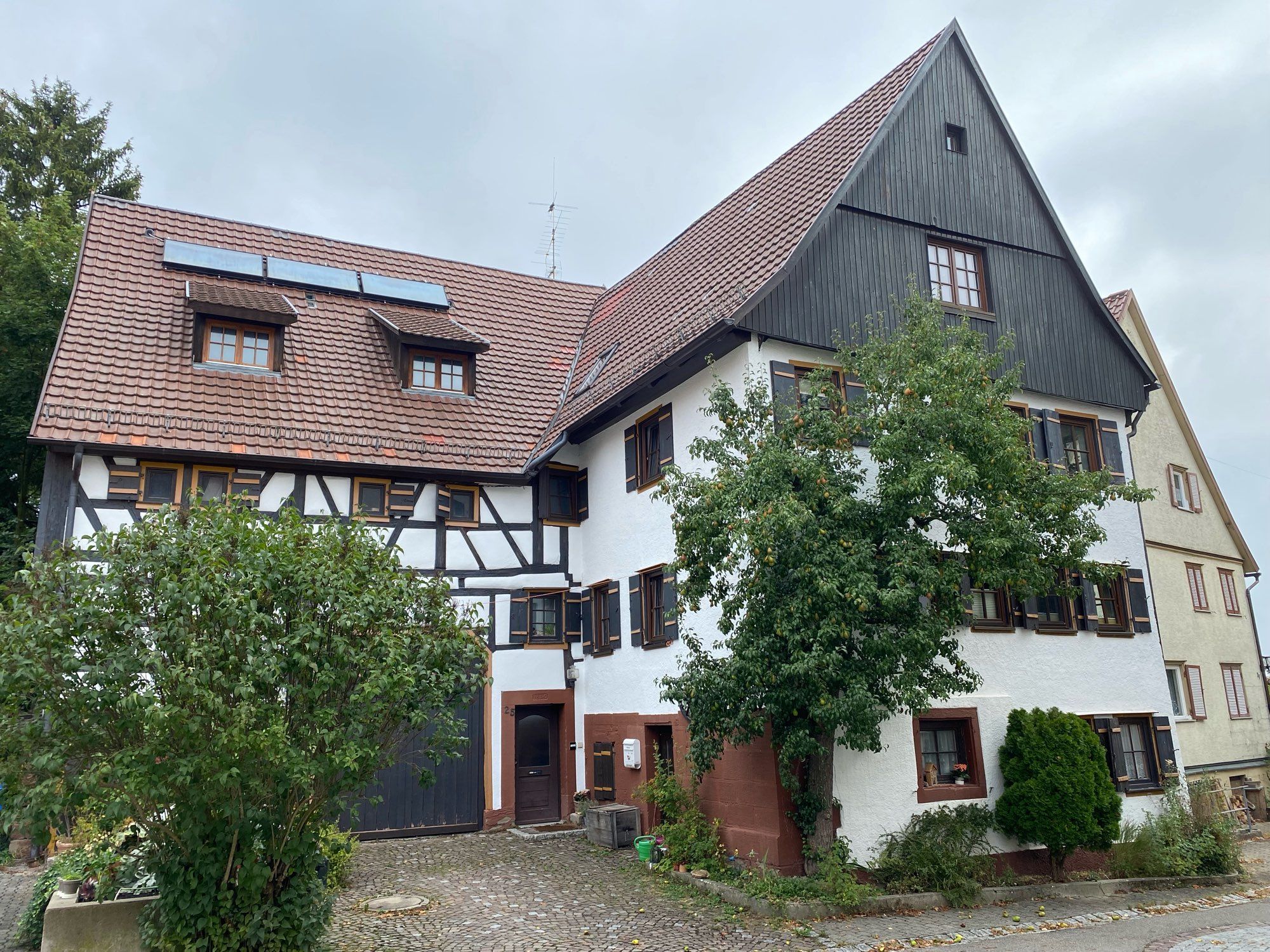 Dreistöckiges, zweiflügliges, altes Bauernhaus. Flügel parallel zur Straße mit Fachwerk und großem Scheunentor. Vorgesetzter Flügel quer zur Straße, mit verputzter Fassade vielen Fenstern mit Fensterläden und holzverkleideter Giebel. Am Eck des Hauses Laubbaum, im gepflasterten Hof zwischen den Flügeln weiterer Laubbaum. Verputzt Wandflächen weiß, Holzelemente dunkelbraun.