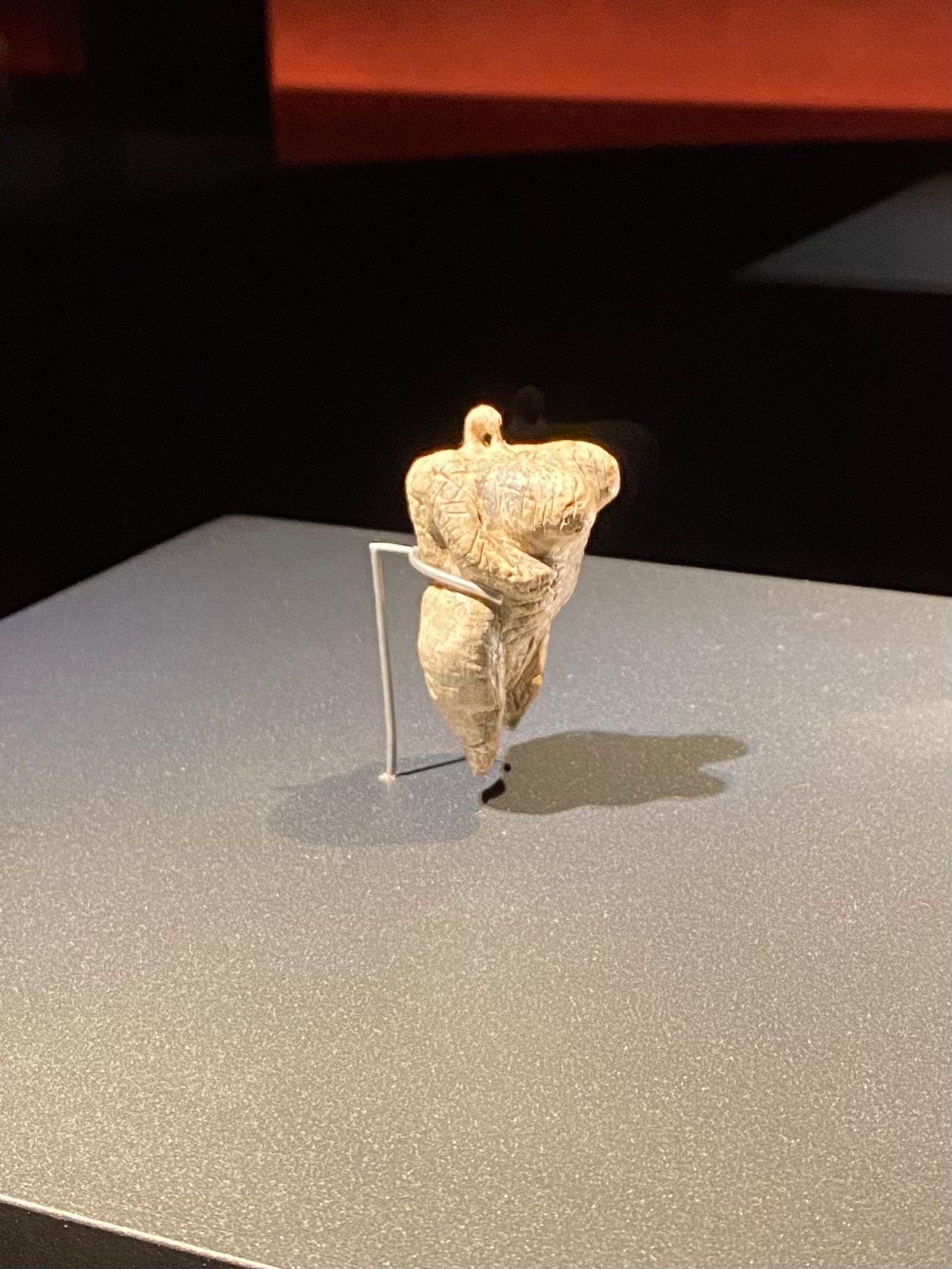 Kleine, aus Elfenbein geschnitzte Skulptur einer menschlichen Figur mit großen Brüsten, Beine kurz und spitz. Statt des Kopfes eine kleine Öse. Von einem Drahtgestell gehalten.