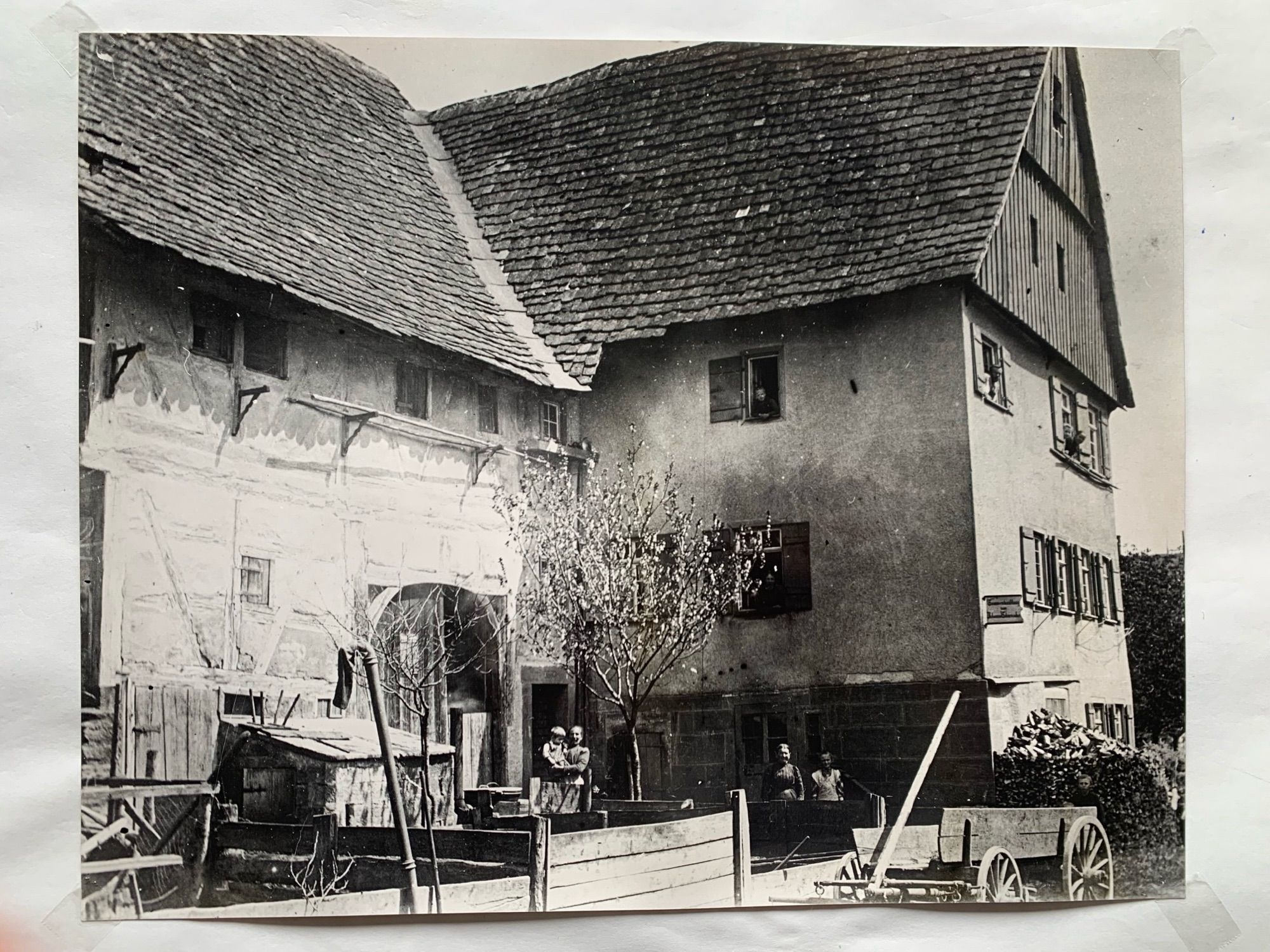 Schwarz-weiß   Dreistöckiges, zweiflügliges, altes Bauernhaus. Flügel parallel zur Straße mit Fachwerk und großem Scheunentor. Vorgesetzter Flügel quer zur Straße, mit verputzter Fassade vielen Fenstern mit Fensterläden und holzverkleideter Giebel. Im Hof Holzgatter von Misthaufen und kleiner Laubbaum. Am Eck des Hauses ein Ladenschild, das nicht lesbar ist. Auf der Straße ein Holzkarren. Im Hof haben sich mehrere Menschen für das Foto aufgestellt. Aus mehreren Fenstern schauen ebenfalls Menschen.