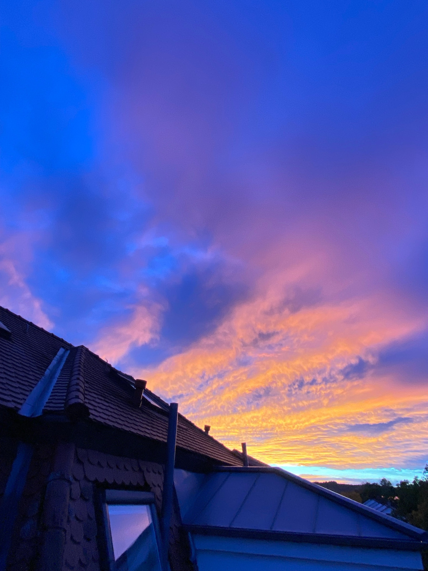 Leuchtend orange-violetter, bewölkter Himmel, als ob von links unten orangene Strahlen über den Himmel färben. Im Vordergrund verwinkeltes Dach mit Biberschwanz-Ziegeln, Dachfenster und Blech-Verwahrungen