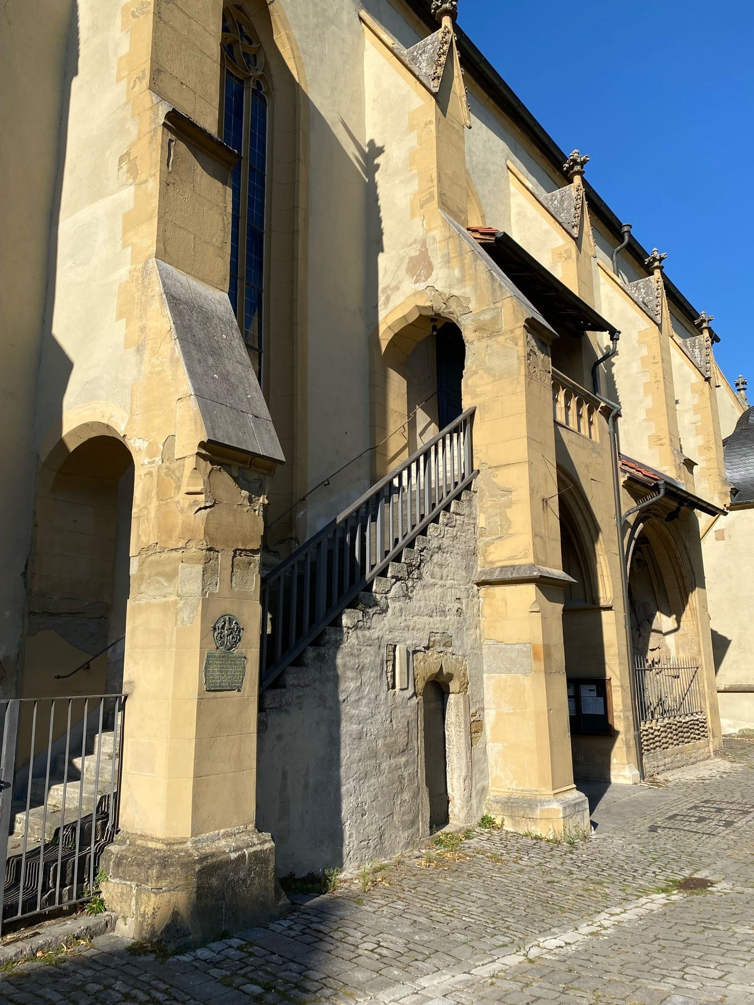 Offener Treppenaufgang außen an einer Kirche aus hellem Stein, die durch Durchbrüche in den seitlichen Wandpfeilern.