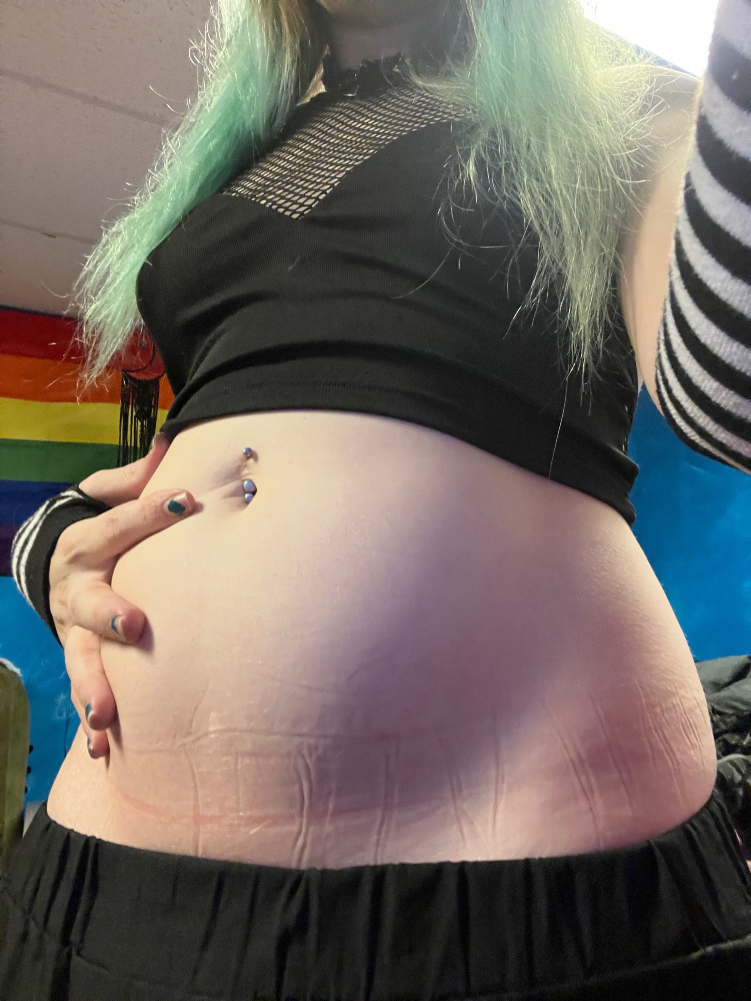 A tummy photo of VaalorieVorse. She’s wearing a black fishnet top, striped arm warmers, and has long blue hair. There is the pride flag in the background against a blue wall.