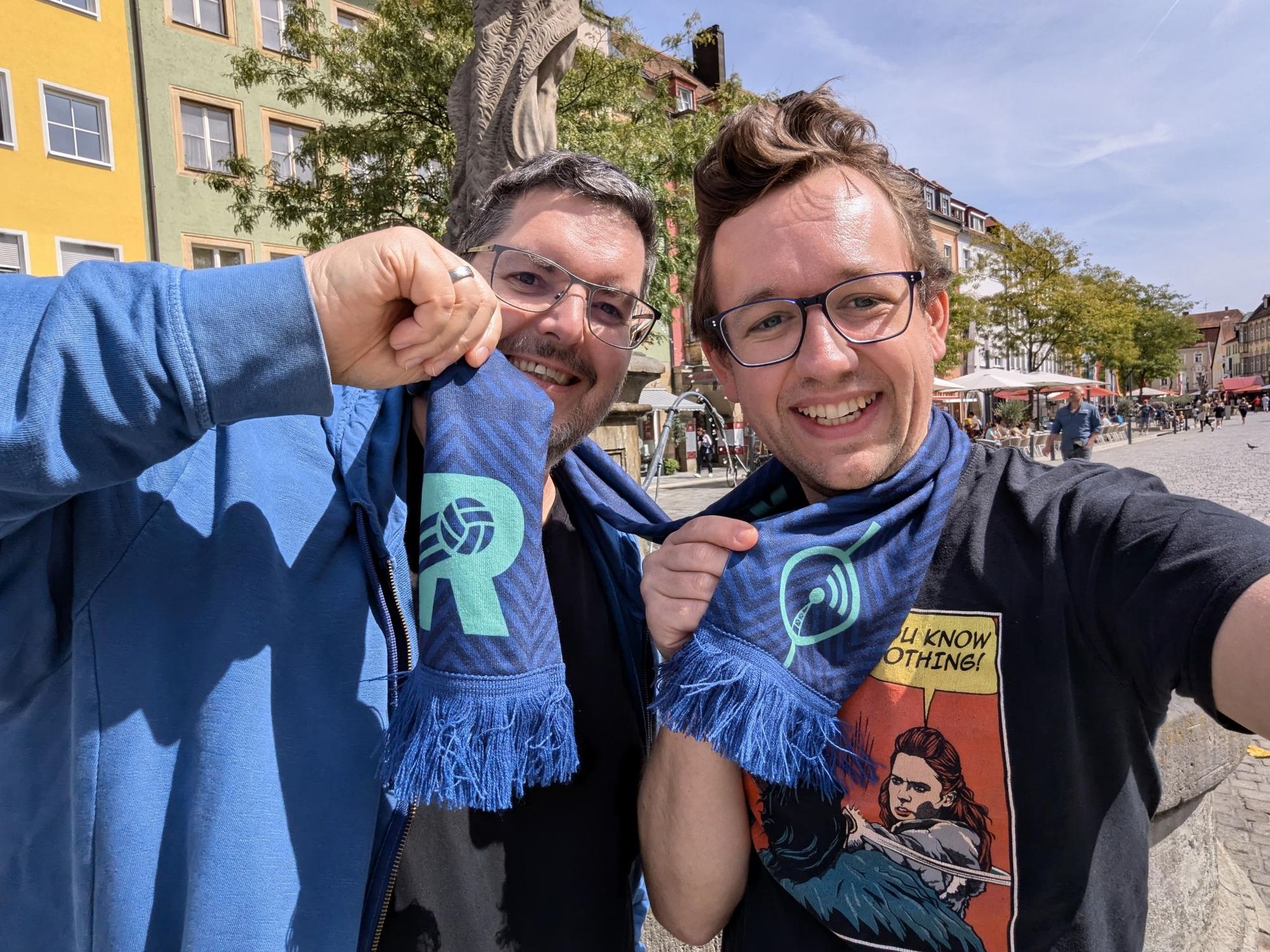 Frank und Max halten einen Schal in der Hand, links mit dem neuen Rasenfunklogo, rechts mit dem alten. Sie sind sehr happy.