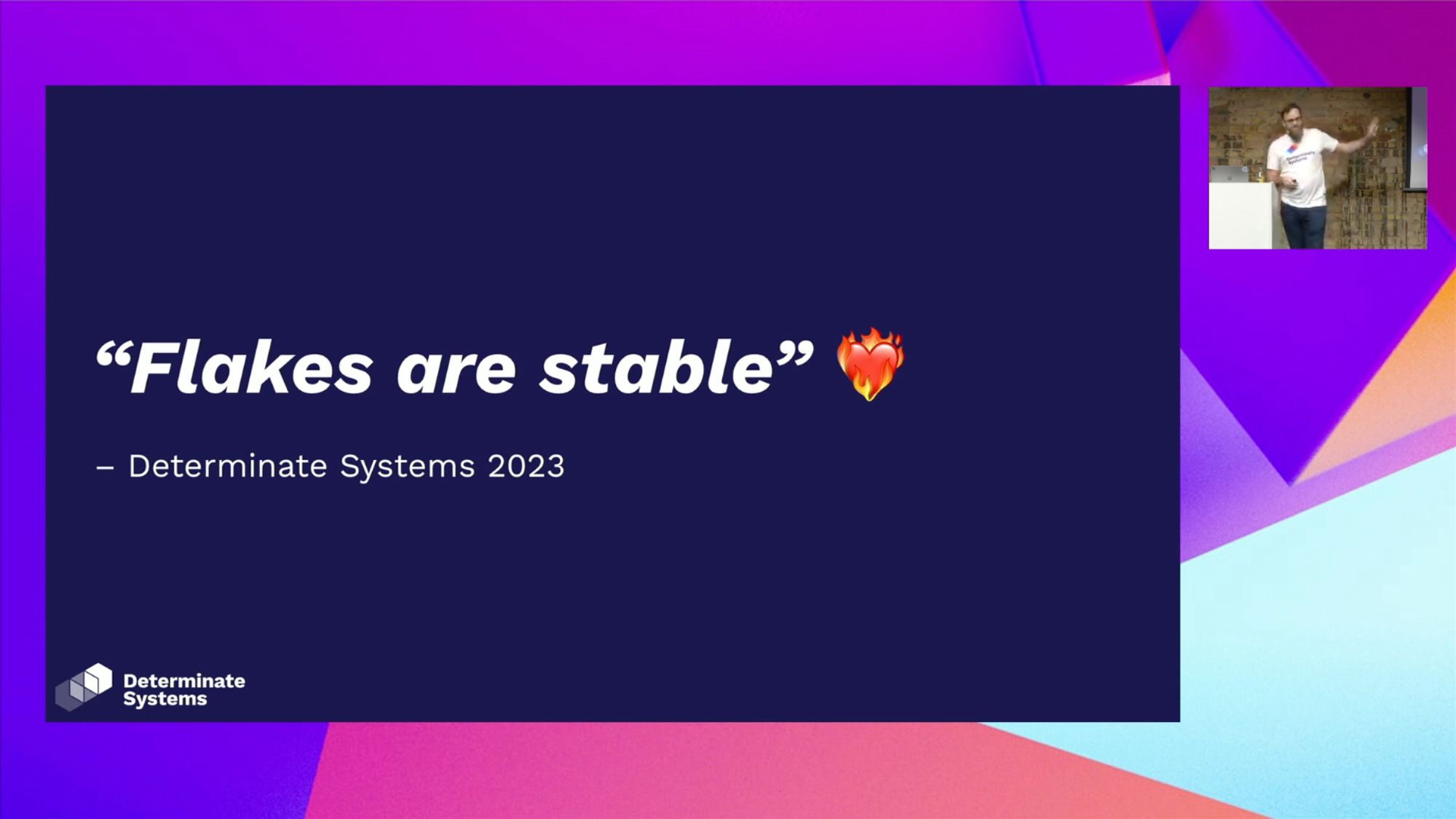 A slide with a dark purple background, and the text has a quote in italics bold "Flakes are stable" with a heart-on-fire emoji. The quote is attributed to Determinate Systems, 2023, since the upstream project has not yet committed to stable Flakes -- but we have. In the corner of the screen is Graham on stage, presenting on the benefits of flakes and why they are the future of Nix.