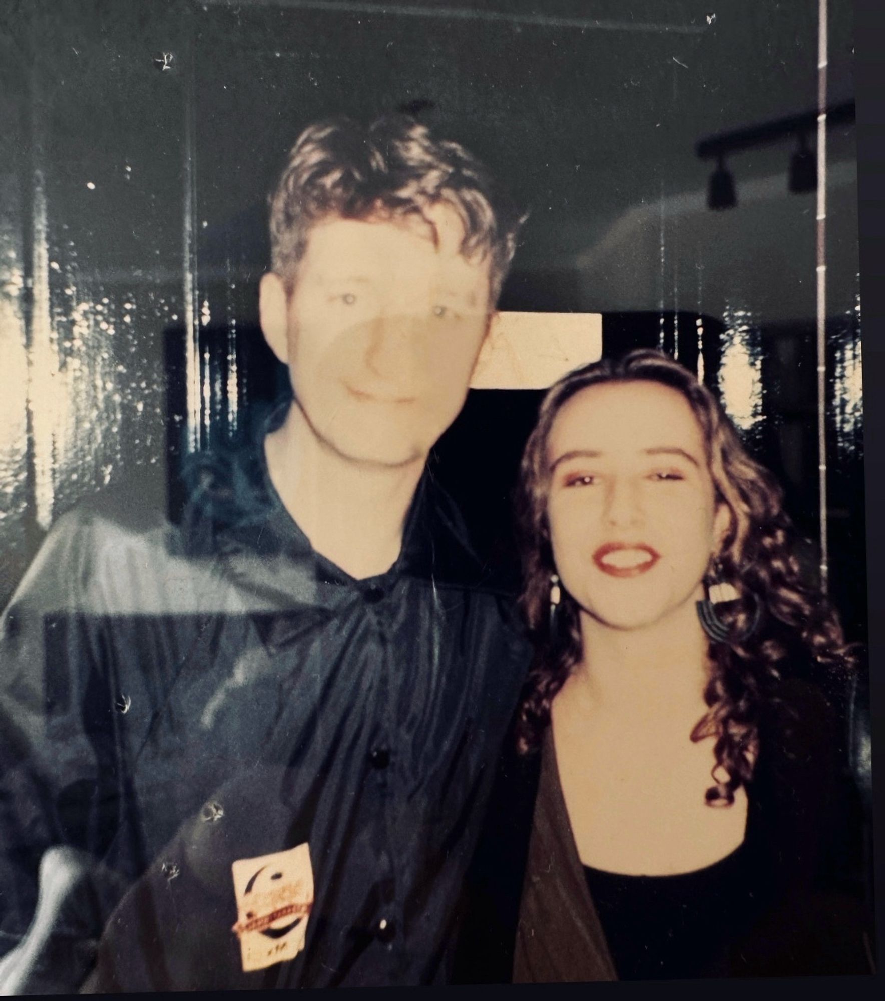 A shot of two people in the photo. From the chests up. On the least is a Caucasian male in his early 30s in a black jacket wsmiling. On the right is a young Caucasian woman with long brown curly hair and red lipstick, smiling.