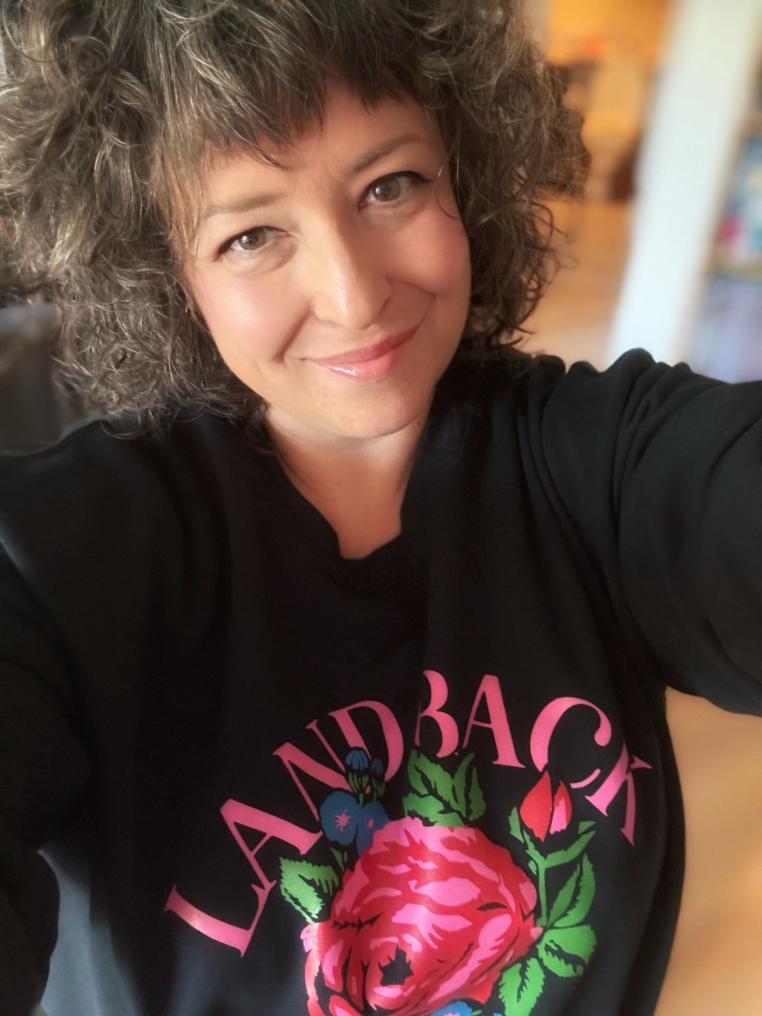 Photo of a middle-aged Caucasian woman with light brown curly hair in a black sweatshirt that says LANDBACK in pink letters with a pink and red rose