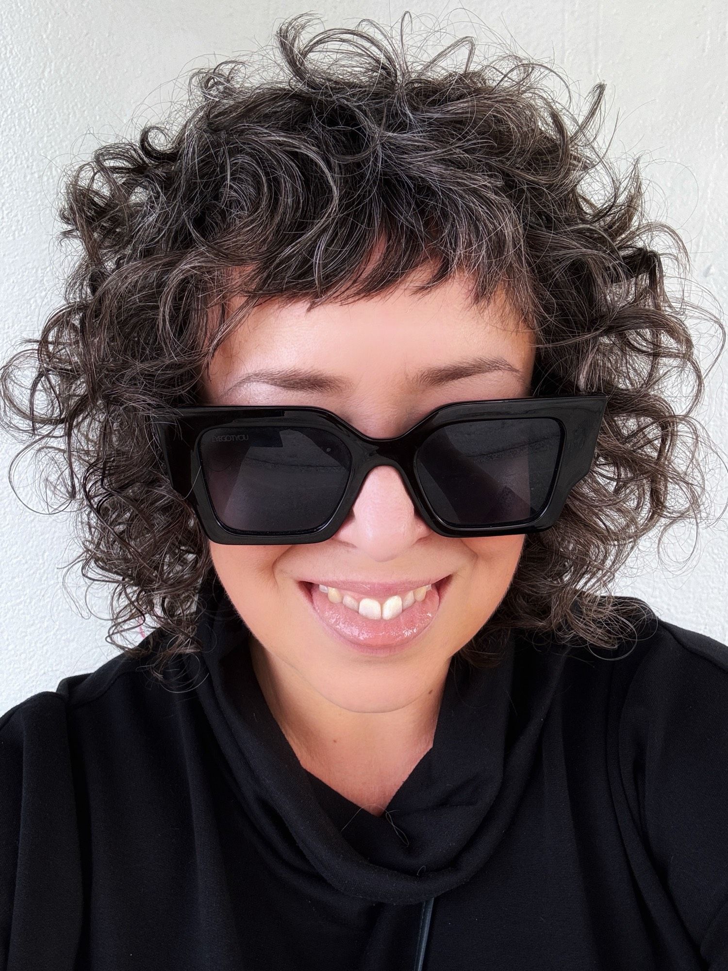 Middle-aged white woman with brown shoulder-length curls and square black si glasses. She is sticking her tongue out