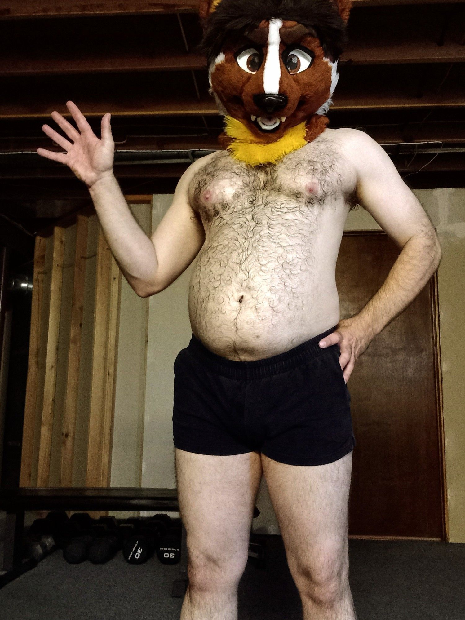 Hairy shirtless man with weasel fursuit head, body relaxed, with his hand waving to the camera.