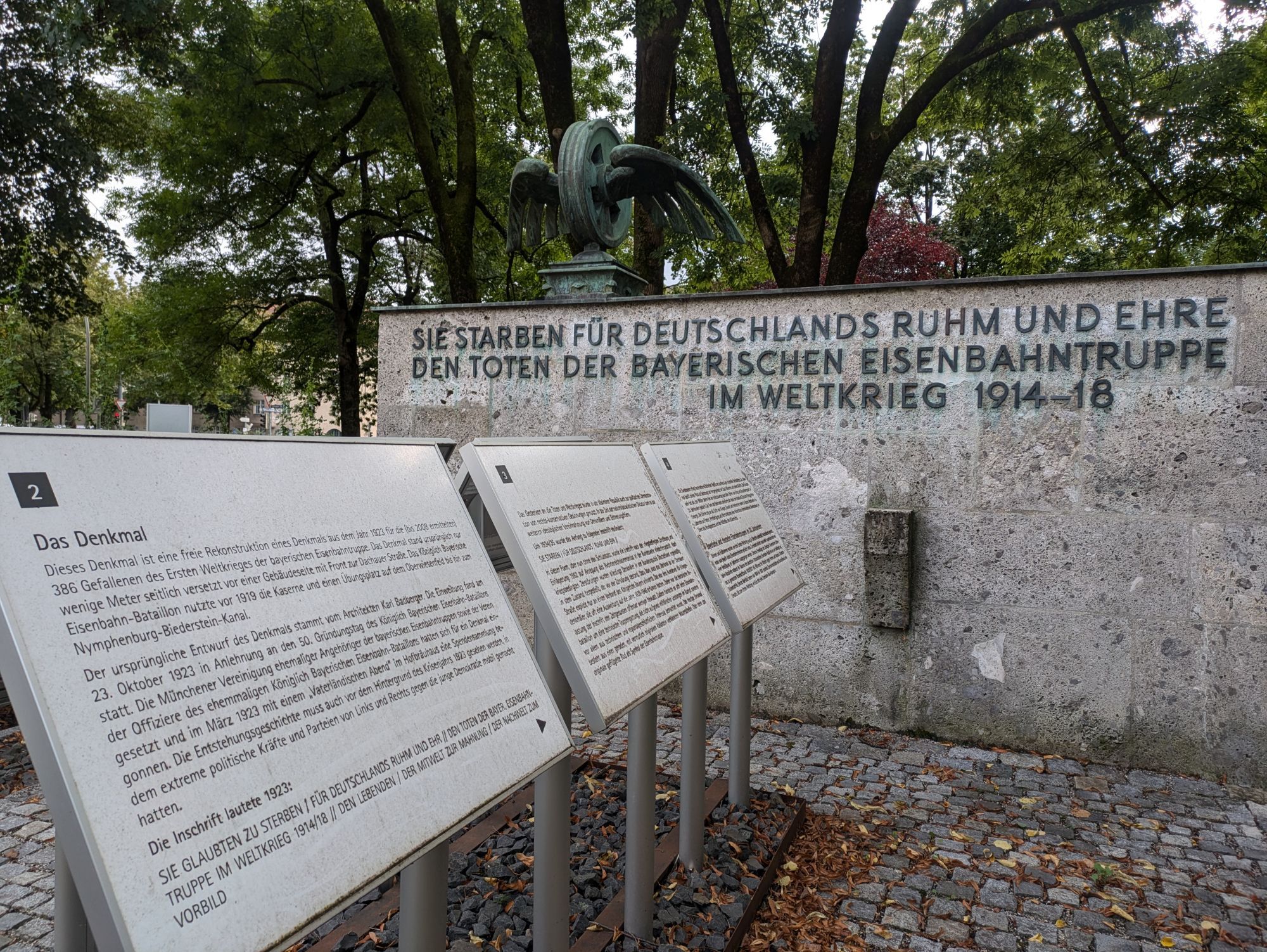 Fabrfoto eines Denkmals mit Geflügeltem Bahnrad aus Metall über Wand mit Inschrift 
"SIE STARBEN FÜR DEUTSCHLANDS RUHM UND EHRE
DEN TOTEN DER BAYERISCHEN EISENBAHNTRUPPE
IM WELTKRIEG 1914-1918"
Davor im Bild 3 von 8 großen weißen Tafeln mit Nummern und Text, lesbar "2 Das Denkmal" und weiter unten 
"Die Inschrift lautete 1923: SIE GLAUBTEN ZU STERBEN FÜR DEUTSCHLANDS RUHM UND EHR (...) DEN LEBENDEN, DER MITWELT ZUR MAHNUNG, DER NACHWELT ZUM VORBILD"