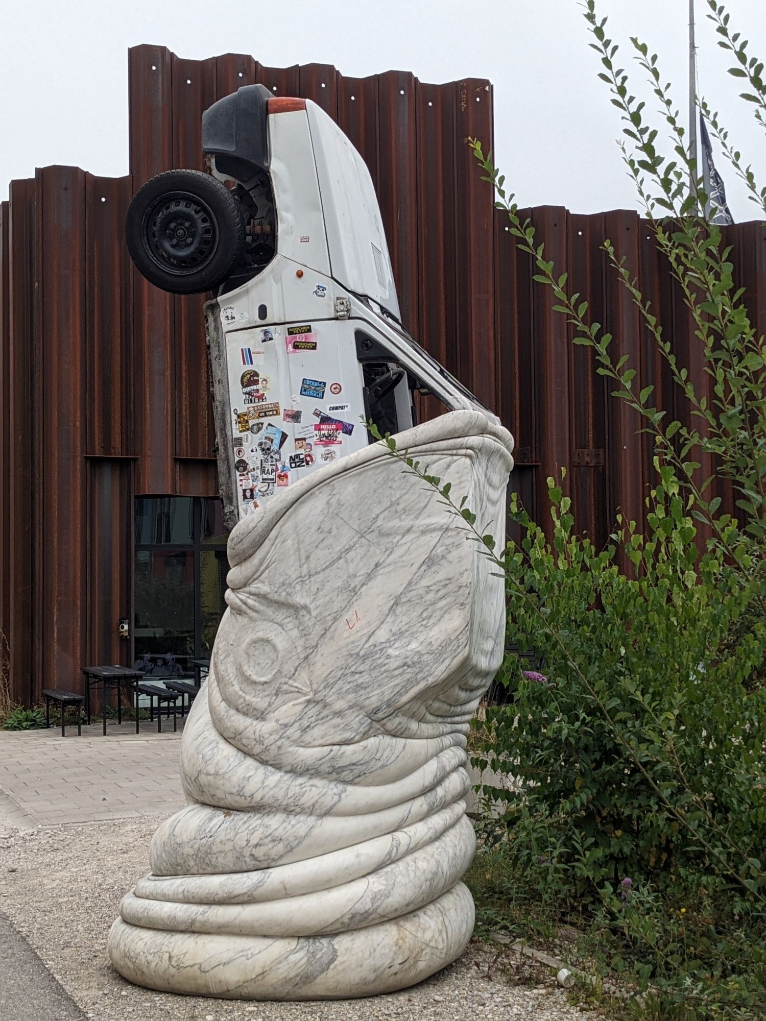 Farbfoto hochkant: Die Skulptur besteht unten aus einem großen fischähnlichen Kopf mit nach oben offenem Maul aus Marmor mit grauer Maserung. Aus dem Maul schaut das obere Ende eines weißen PKW heraus, dessen Seiten mit verschiedenfarbigen Aufklebern bedeckt sind.
Die Skulptur steht auf Asphalt neben einem Strauch vor einem Gebäude, dessen Fassade aus rostbezogenen Spundwand-Teilen, es stehen dunkle Tische und Bänke davor. Der Himmel oben ist undefinierbar grau.