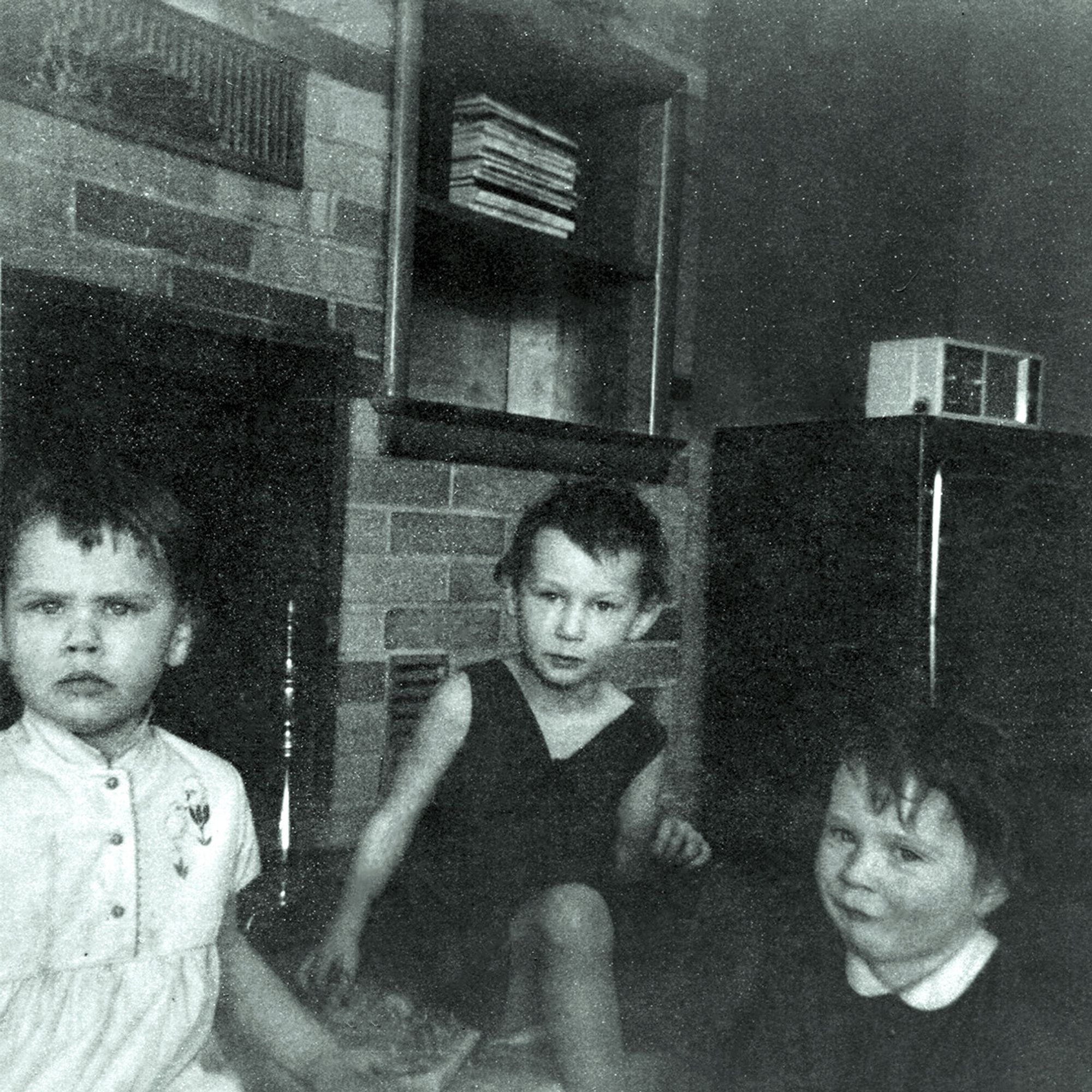 Black and white photo of three girls under five years old sitting on a hearth with a television in the background.