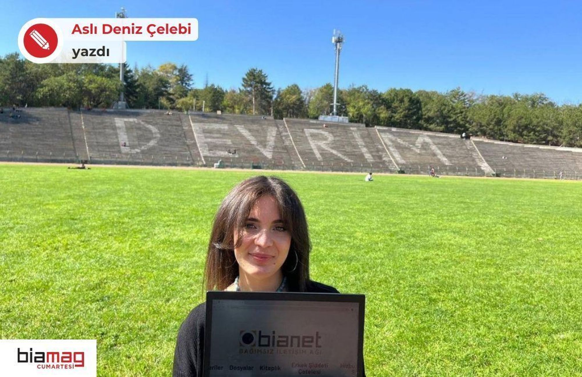 Fotoğrafta, geniş bir yeşil alanın önünde bir kadın duruyor. Kadın, elinde "Bianet Bağımsız İletişim Ağı" yazılı bir tablet tutuyor. Arka planda büyük bir stadyum tribünü görülüyor ve tribün sıralarının üzerine büyük harflerle "DEVRİM" yazılmış. Hava açık ve güneşli, gökyüzü masmavi. Tribünlerin arkasında ağaçlar yer alıyor, ve stadyumda birkaç kişi uzakta oturuyor. Kadının saçları açık ve hafif gülümseyen bir ifadesi var. Fotoğraf, açık bir alanda güzel bir sonbahar gününü yansıtıyor.