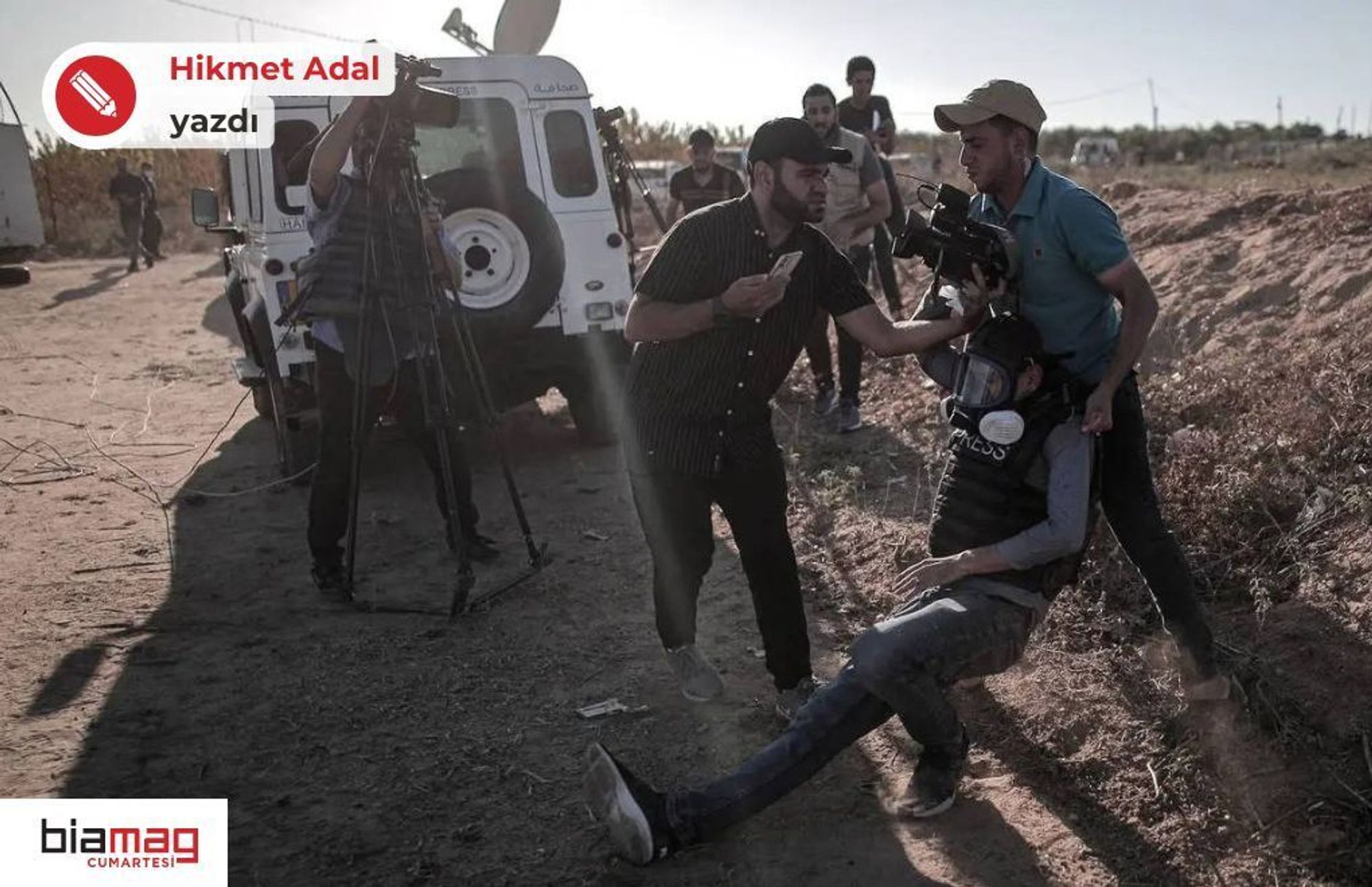 Fotoğrafta bir açık hava sahnesi görülüyor. Görüntüde birkaç kişi yer alıyor ve bu kişiler muhtemelen gazeteciler veya medya çalışanları. Ön planda, yere oturmuş olan bir kişi var; üzerinde "PRESS" (basın) yazılı bir yelek ve miğfer bulunuyor. Bu kişi muhtemelen yaralanmış, çünkü iki kişi onu kaldırmaya veya destek olmaya çalışıyor. Bir kişi miğferine tutunarak destek sağlıyor, diğer kişi ise yanından telefonla yardım çağırıyor gibi görünüyor.  Arka planda beyaz bir araç var, üzerinde de "PRESS" yazısı göze çarpıyor. Araç, bir medya aracı gibi görünüyor ve yanında kamera ekipmanları olan başka kişiler de var. Fotoğraftaki ortam toprak bir arazi, çalılar ve yol kenarına benzer bir yerleşimle çevrili. Işık, gün batımına yakın bir vakitte çekilmiş olduğunu düşündürüyor, çünkü gölgeler uzun ve ışık açısı düşük. Çevredeki hava gergin, acil bir durum yaşanıyor gibi görünüyor.