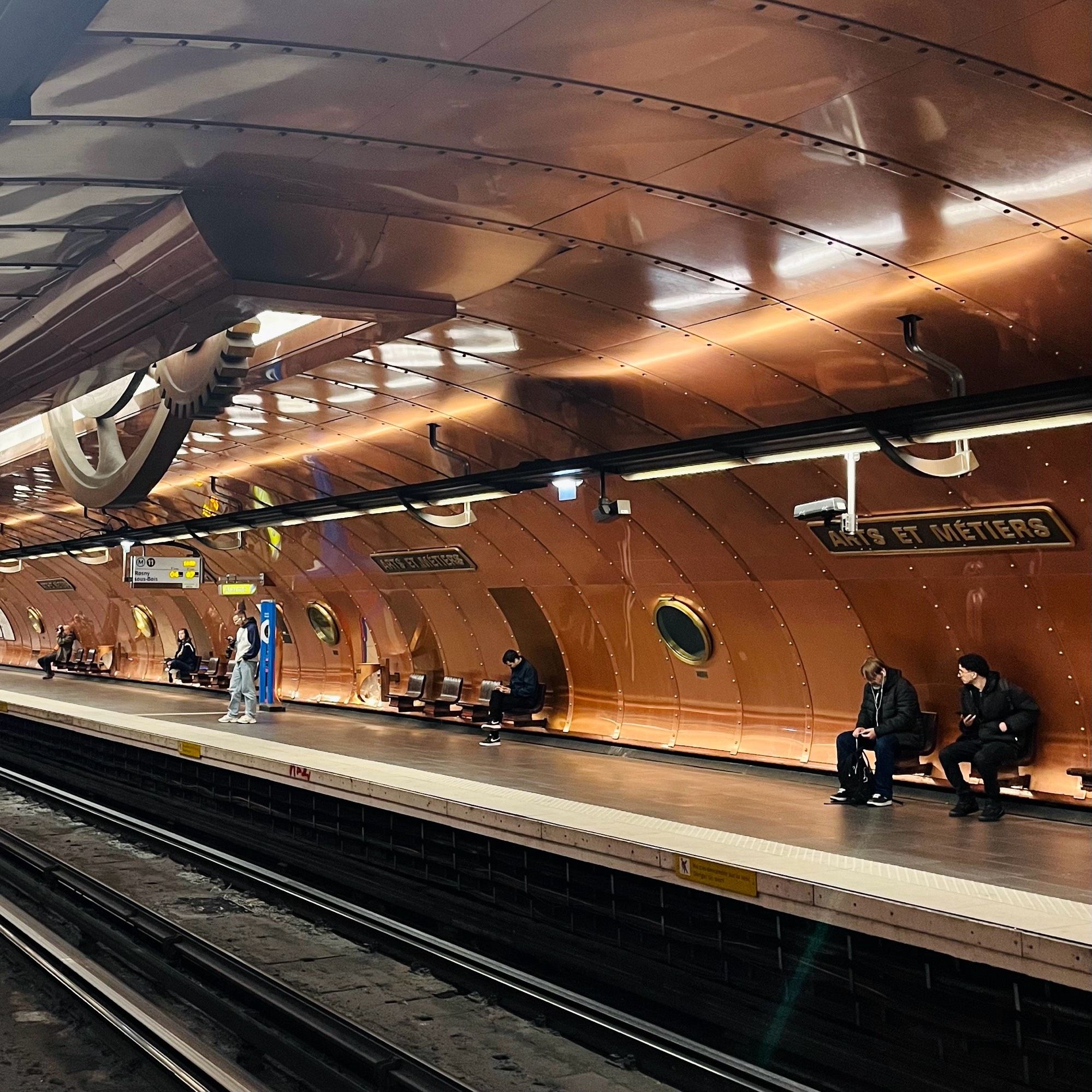 A platform at Arts et Métiers metro station in Paris. The wall is lined with curved panels of copper-coloured metal and there are big cogs coming down from the roof. It looks like you’re trapped in a steampunk machine.