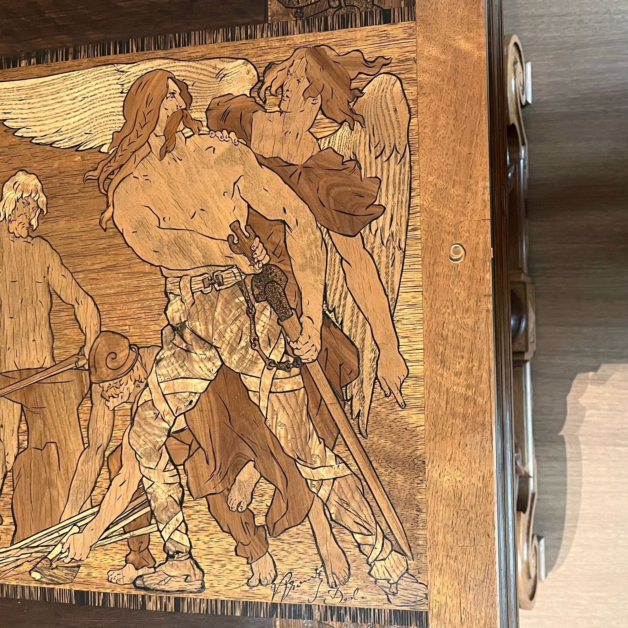 Detail of an Art Nouveau table decorated with a frieze of images created using inlaid wood of different shades. The central figure is a muscular Gaul warrior, who is shirtless qnd carrying a big sword. His hair is long and wavy and so is his moustache. A lady angel is clasping him for her own very good reasons.