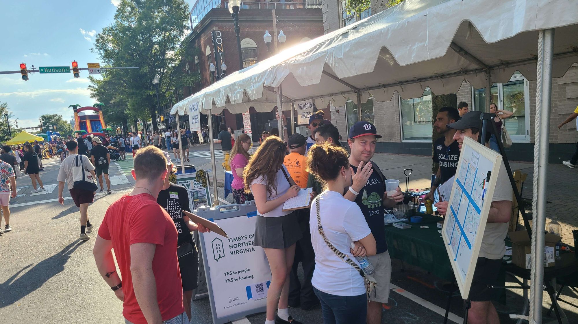 A small group of half a dozen people standing in front of a tent talking and listening. The tent has a sign saying "YIMBYs of Northern Virginia". It is daytime with sunny weather. The booth is at the Clarendon Day festival in Arlington Virginia.