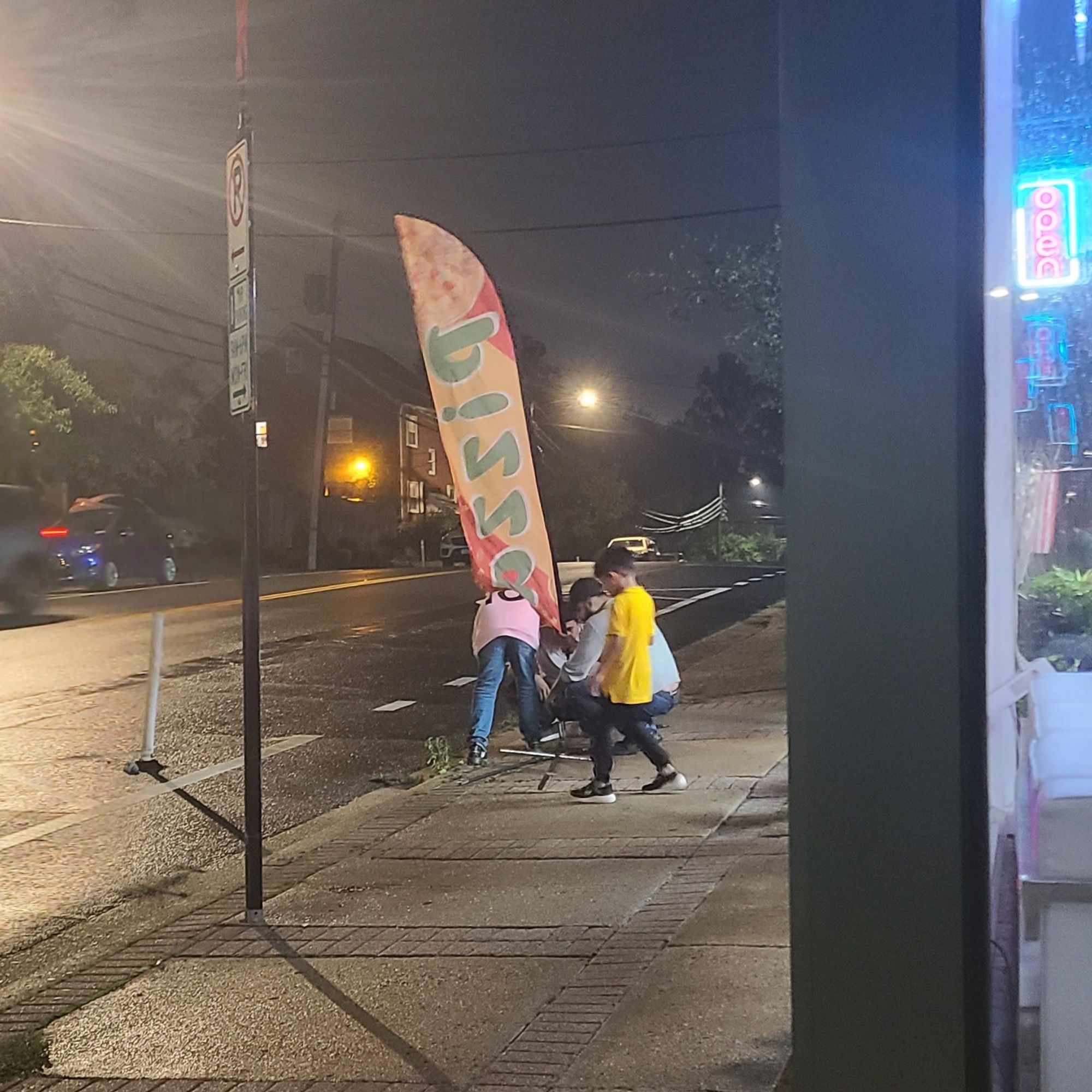 A picture at night of a couple of adults surrounded by a couple of children on a sidewalk crouching down digging a small hole out of a sidewalk. A 6 ft feather flag reading "pizza" is being readied to be put down into it.