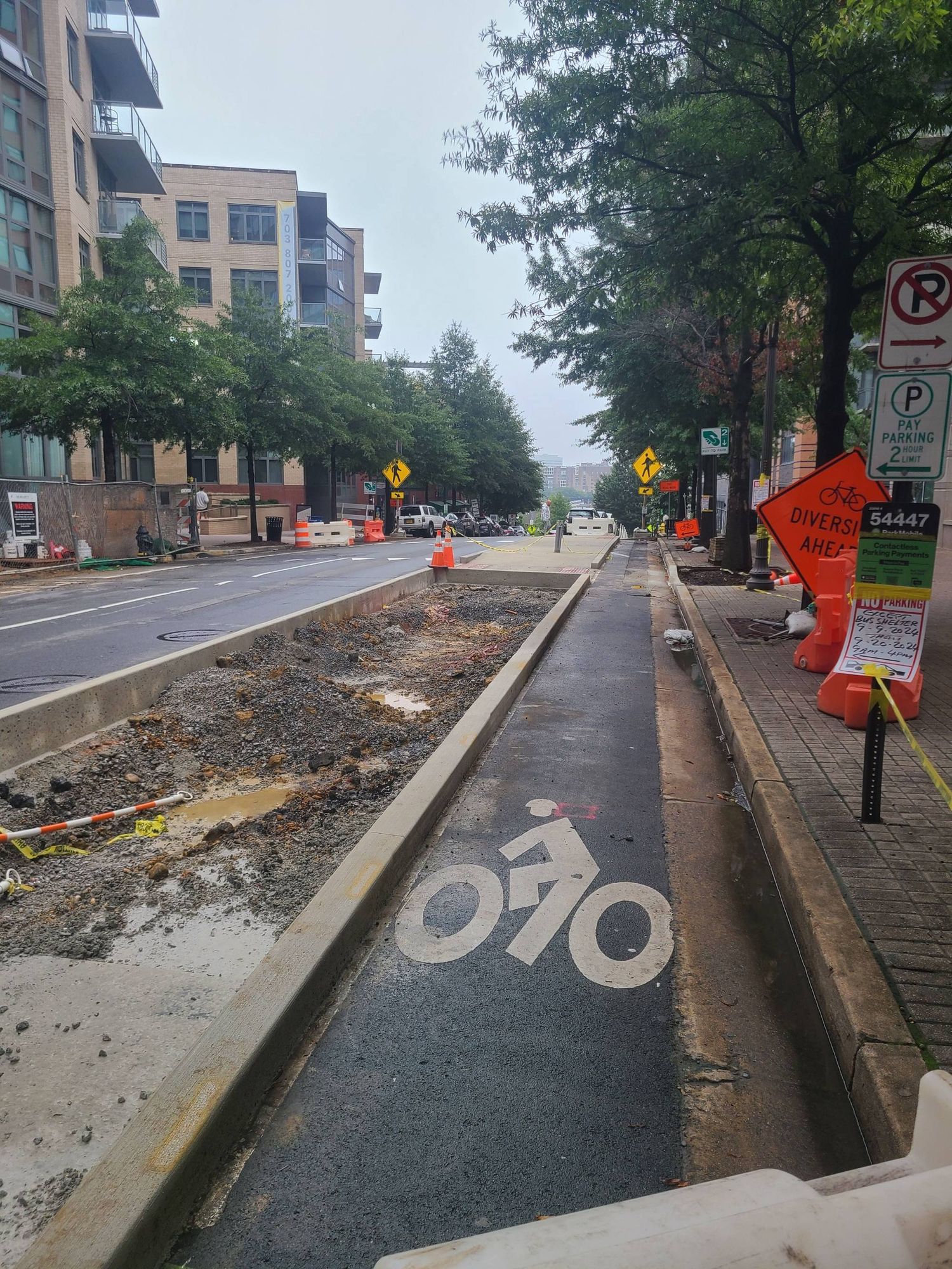 Picture of a city street surrounded by mid-rise buildings there is a protected bike lane under construction separated from the street by a cement tree planter and a bus island.