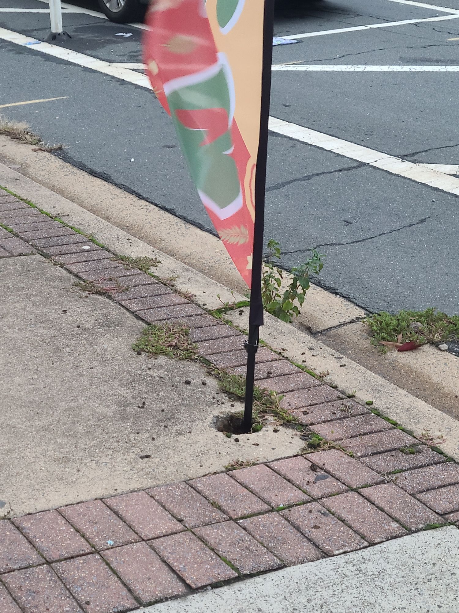 Picture during the daytime of a sidewalk with a 3-in hole dug into it and a flagpost inserted into it.