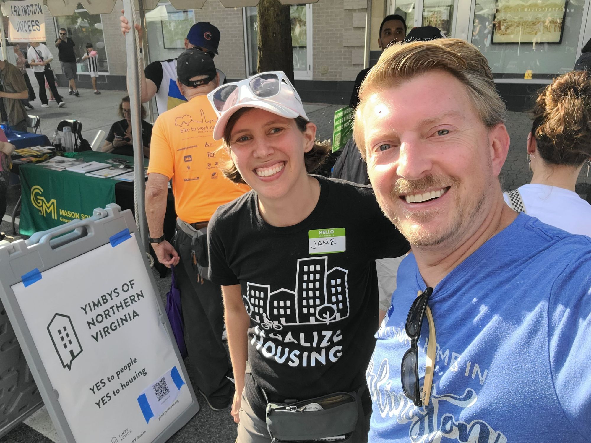 Picture of a man and a woman in pro-housing related shirts smiling to the camera and standing in front of a sign stating "YIMBY's of Northern Virginia. Yes to people. yes to housing". The booth is at Clarendon Day in Arlington Virginia.