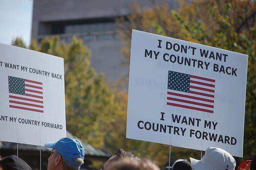 Protesters holding white signs with USA flag in middle and black text reading, "I don't want my country back. I want my country FORWARD!"