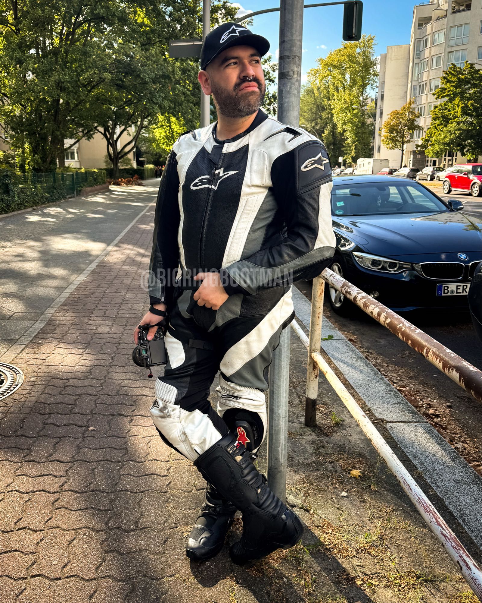 Man wearing an Alpinestars riding suit and boots. Matching hat, and holding a camera.