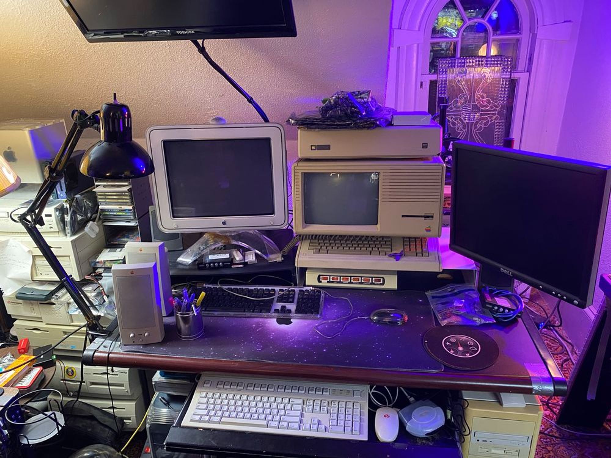 Computer desk setup with a shelf full of Apple Macintoshes to the left. On the desk is an Apple CRT display next to a vintage Apple Lisa that is sitting roughly in the middle. To the right of the Lisa is a 4:3 ratio LCD display mounted to a monitor arm. 