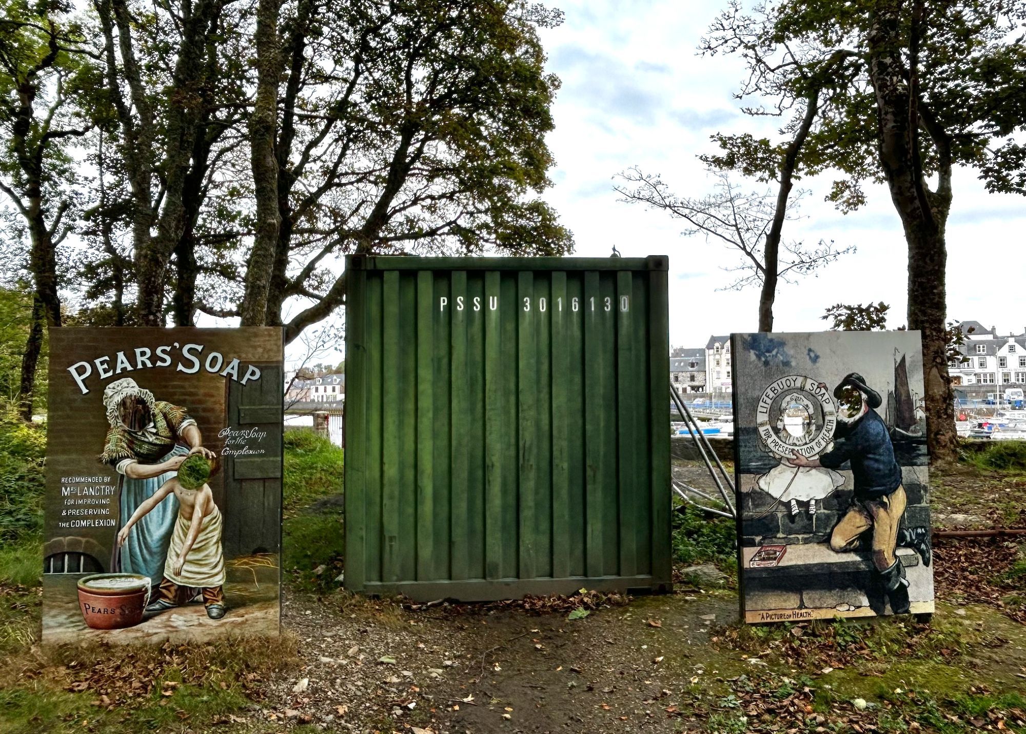 a large green bin positioned between two retro soap ads, each with cut outs for people to put their faces in. various metal bars and castaway items are behind them, rendering them odd tourism items