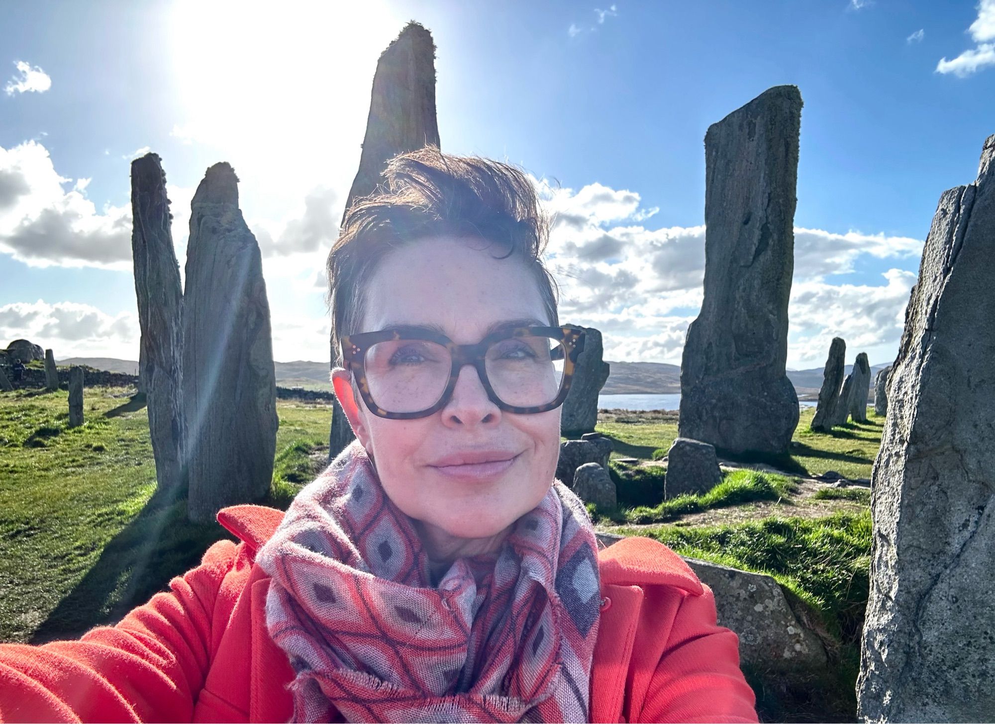 white femme person in short hair and glasses, with scarf and orange coat, in front of ancient standing stones