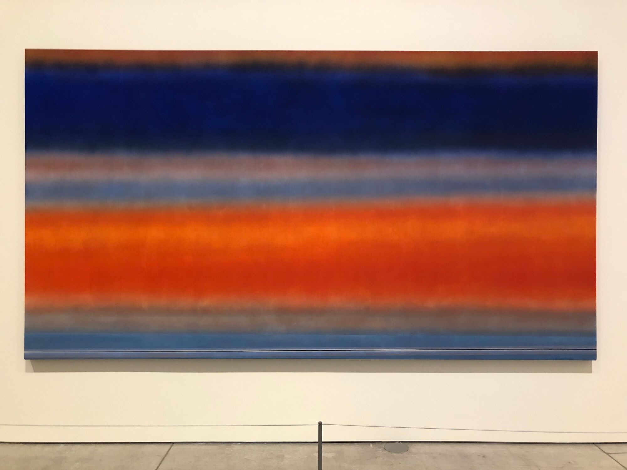 Daybreak, by Rita Letendre, 1983
Acrylic on canvas

Dark navy blue, thick, blurry, horizontal line, grey and light blue blurry lines much thinner below it, thick, bright deep orange blurry line below those, another grey and light blue thinner blurry lines under that. Your eyes try to focus but can’t and it creates an illusion of movement from the navy blue to the orange and back and forth. 

From title plate on wall:
During the 1970s, many artists, including Letendre, began working with airbrushes. The tool allowed the artist to control her paint application, resulting in pure, flat planes of colour. In Aforim, she used an airbrush to soften hard edges in the upper and lower sections of the work. The atmospheric fields of colour in Daybreak highlight the evolution of Letendre’s airbrush technique.