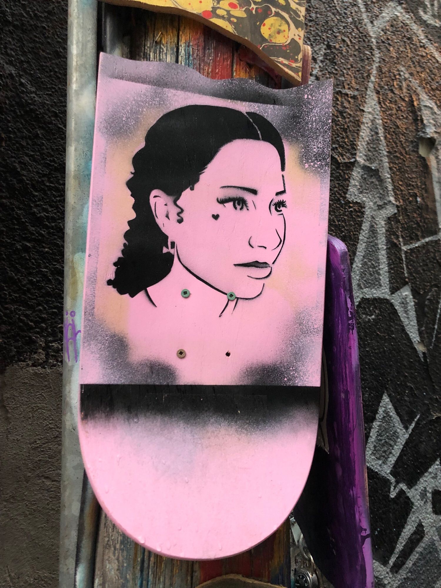 A black stencil of a woman’s face on pink background, 3/4 side profile, black wavy hair pulled back and heart tattoo on her upper cheek near her eye.