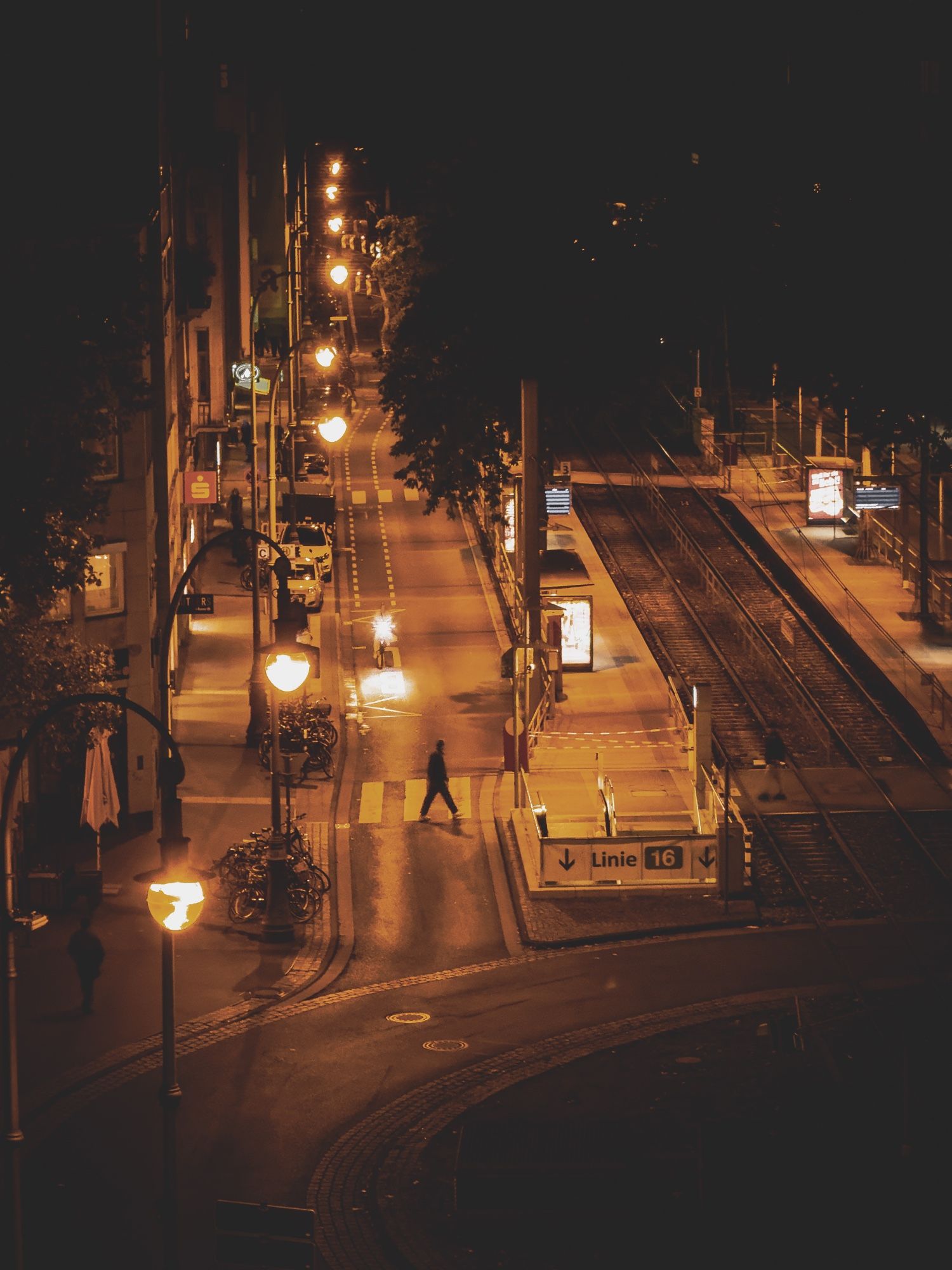 A photo of a street at night from several stories above, with orange streetlights leading through the frame, illuminating a cyclist and a pedestrian.