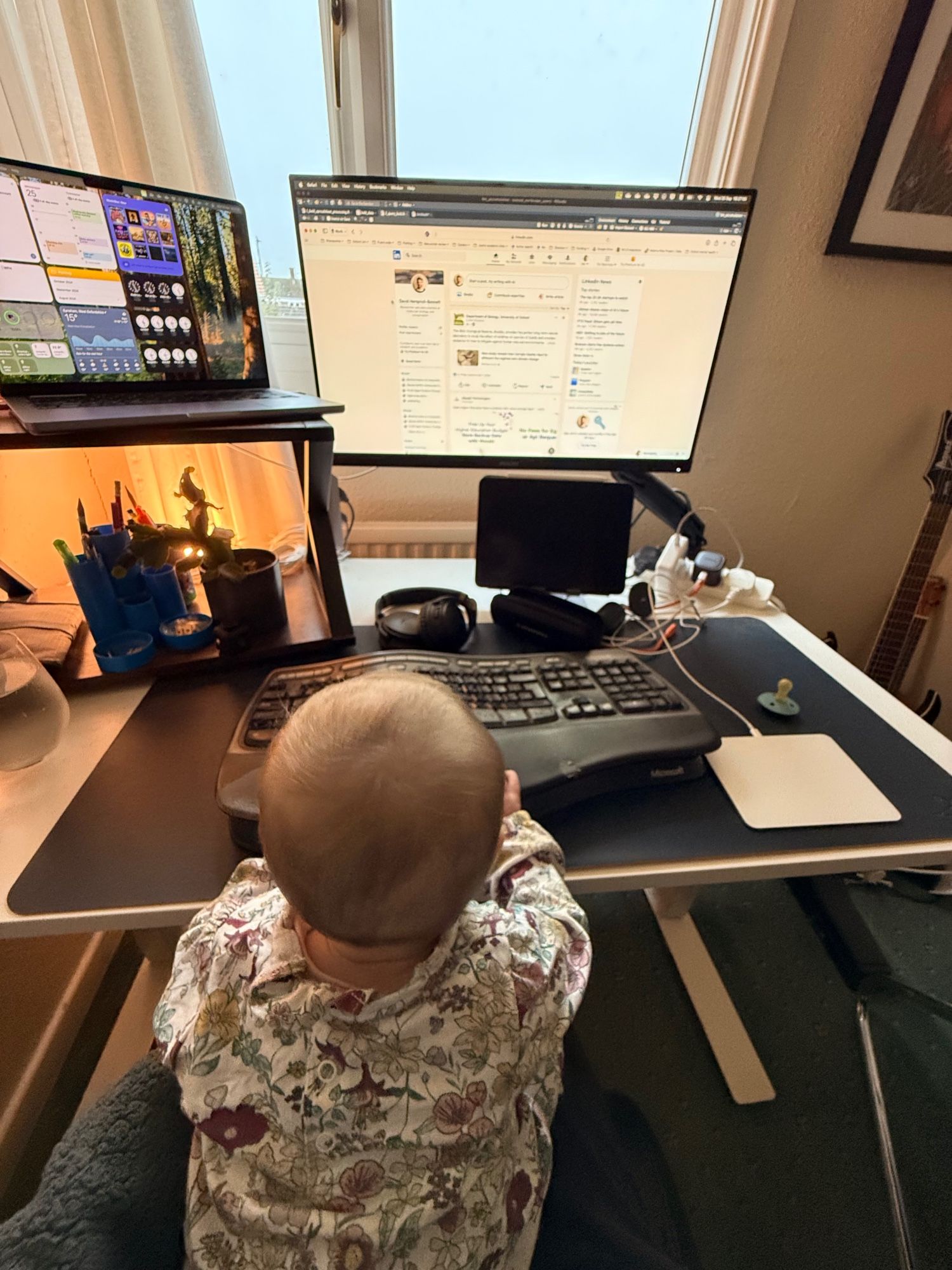 Photo of a small baby sat at a computer desk, pressing a keyboard, with LinkedIn open onscreen and a dummy next to the trackpad