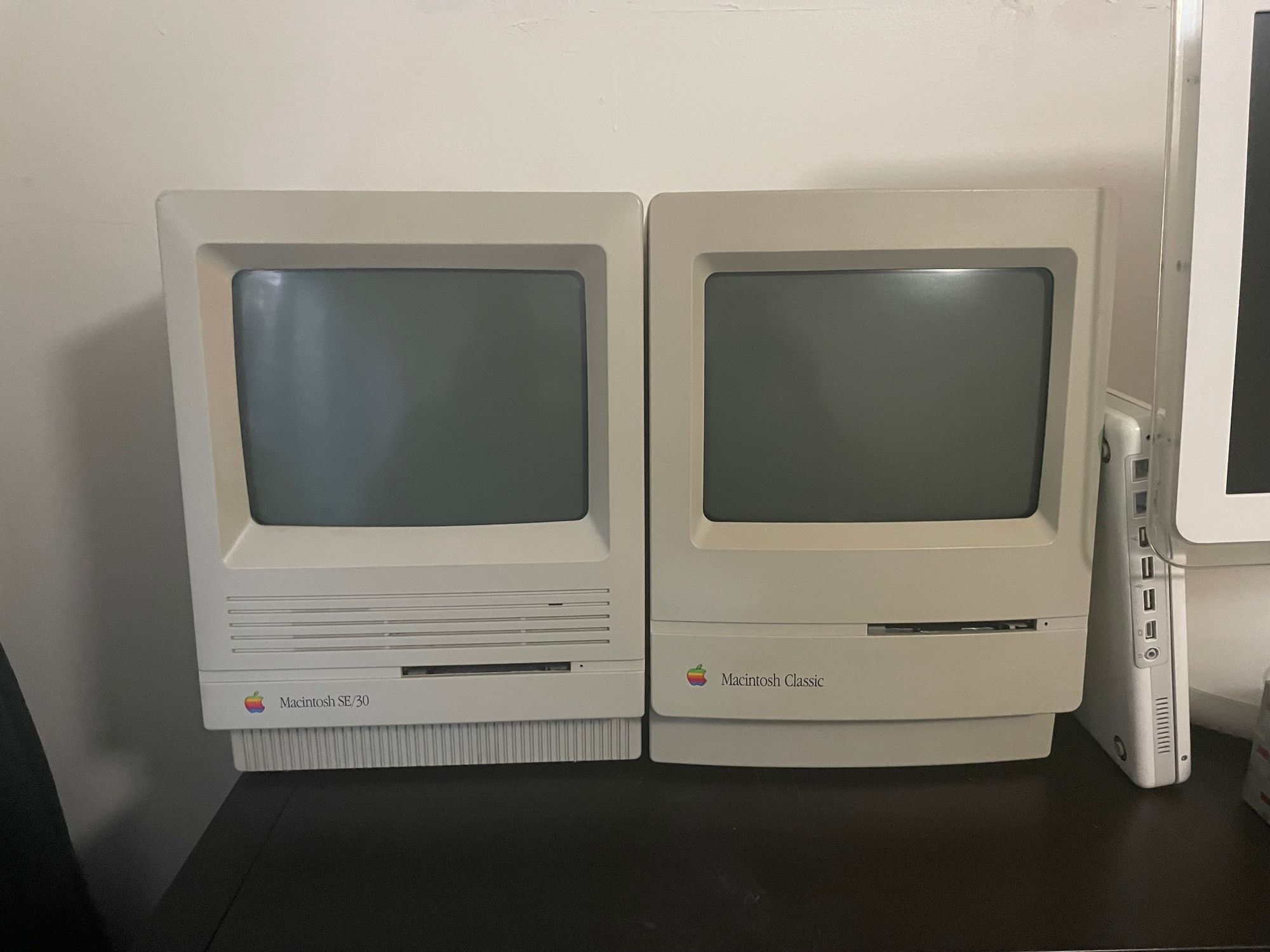 A photo of two compact early 90’s Macintosh computers next to each other on my black shelf. On the left is a Macintosh SE/30, and on the right is a Macintosh Classic. 

Further to the right you can see the side ports on a propped-up white iBook G3, and the edge of an iMac G4 LCD.