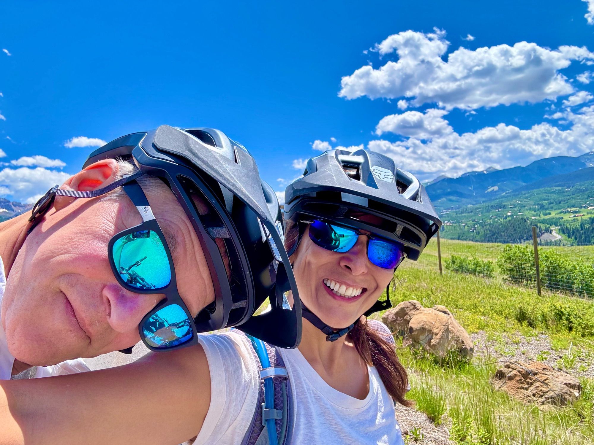 Closeup of me and my husband in bike helmets and sunglasses, with green hills & blue mountains in the background, blue skies and clouds.