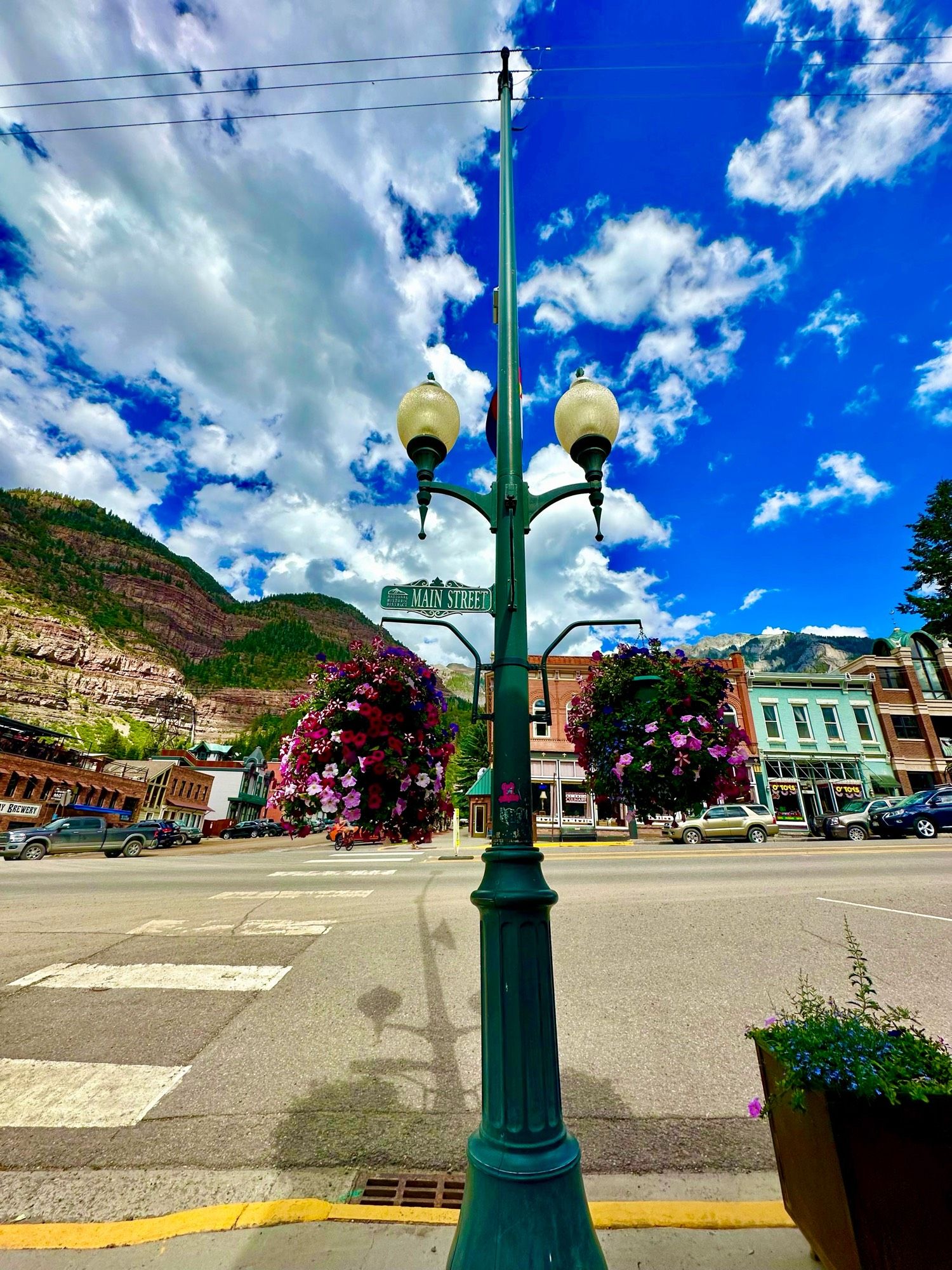 View looking across a Main Street, with an ornate street lamp with two flower baskets in foreground, mountains in background with blue sky and clouds.