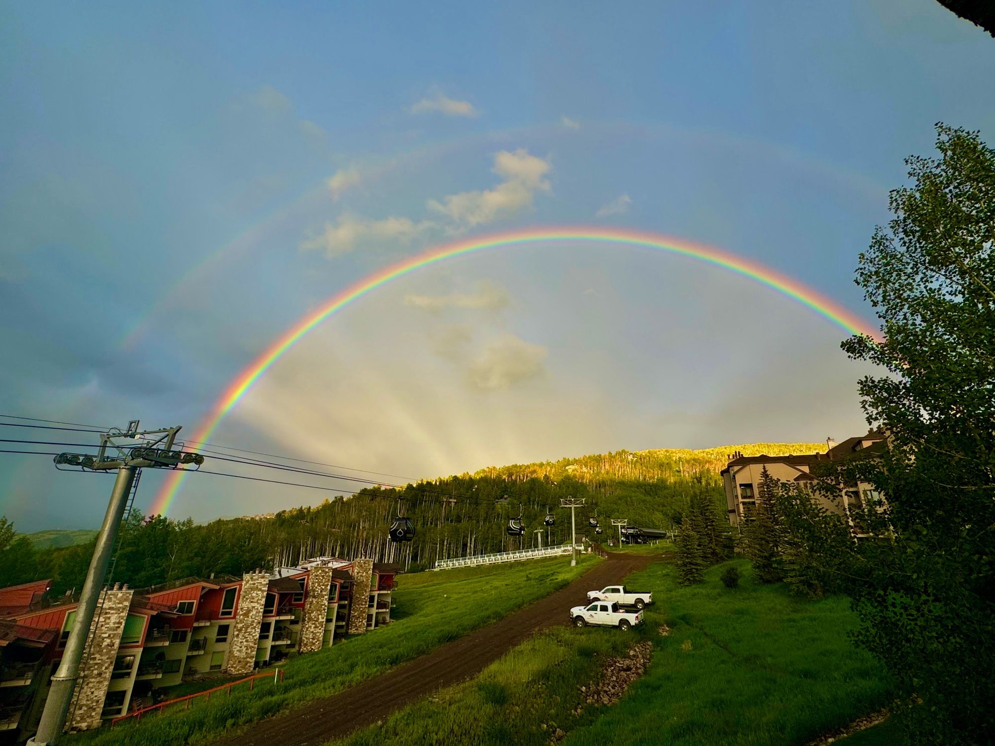 A double rainbow spans the width of the photo above a trees and some buildings to the left.