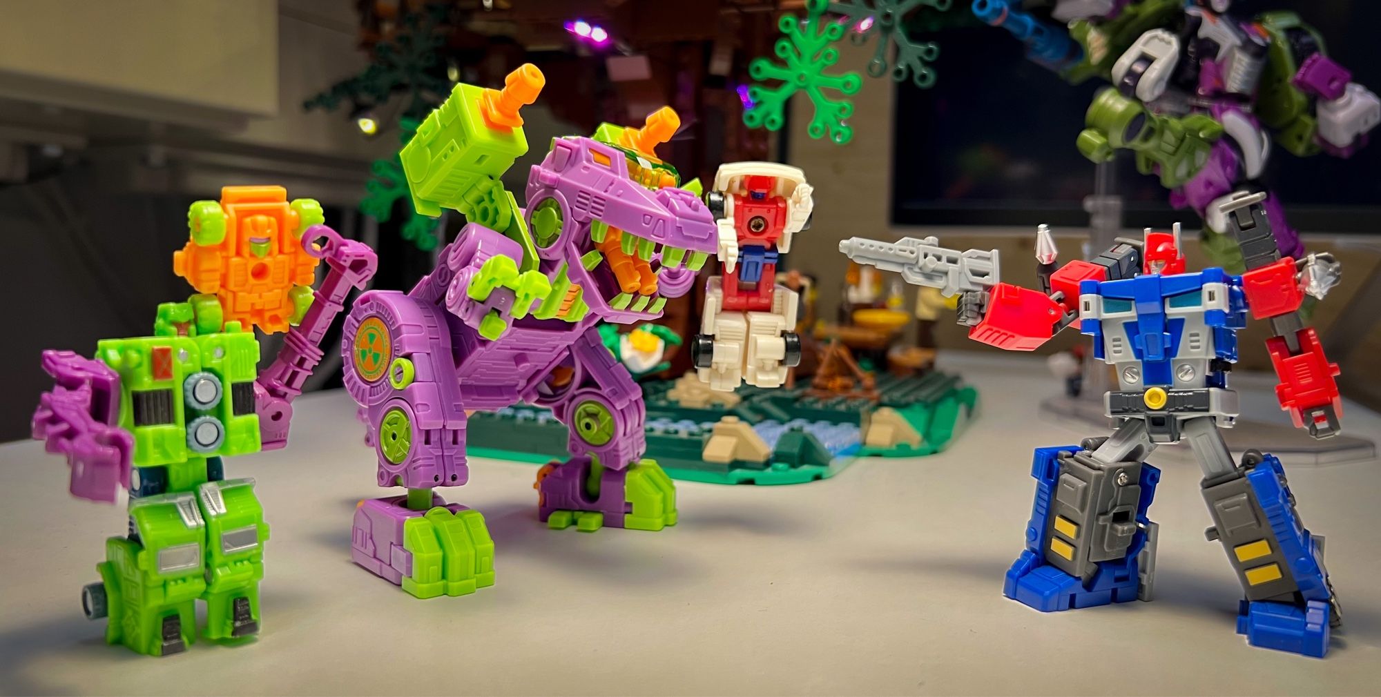 The base has turned into the garishly colored robot dinosaur Trypticon, who is holding poor fixit in one of his claws. Full-Tilt is standing on the shoulder of a construction vehicle Minicon and looking in at the side of Trypticon, while Delta Magnus, now in bot mode, is pointing his blaster towards the robo dinosaur.