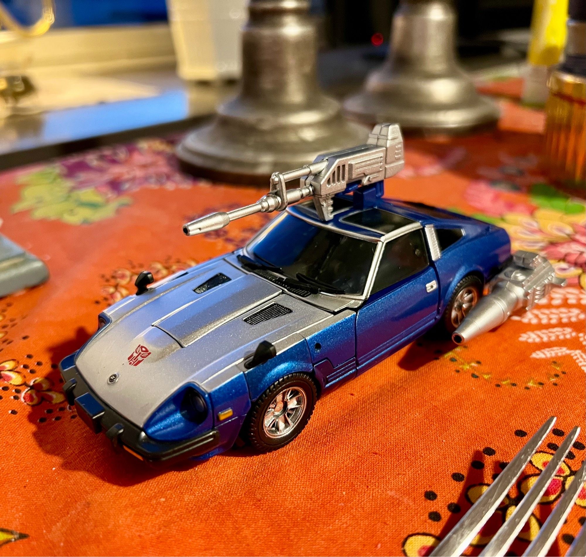 Masterpiece Blue Bluestreak in vehicle mode, all guns attached, next to a fork on a dining room table