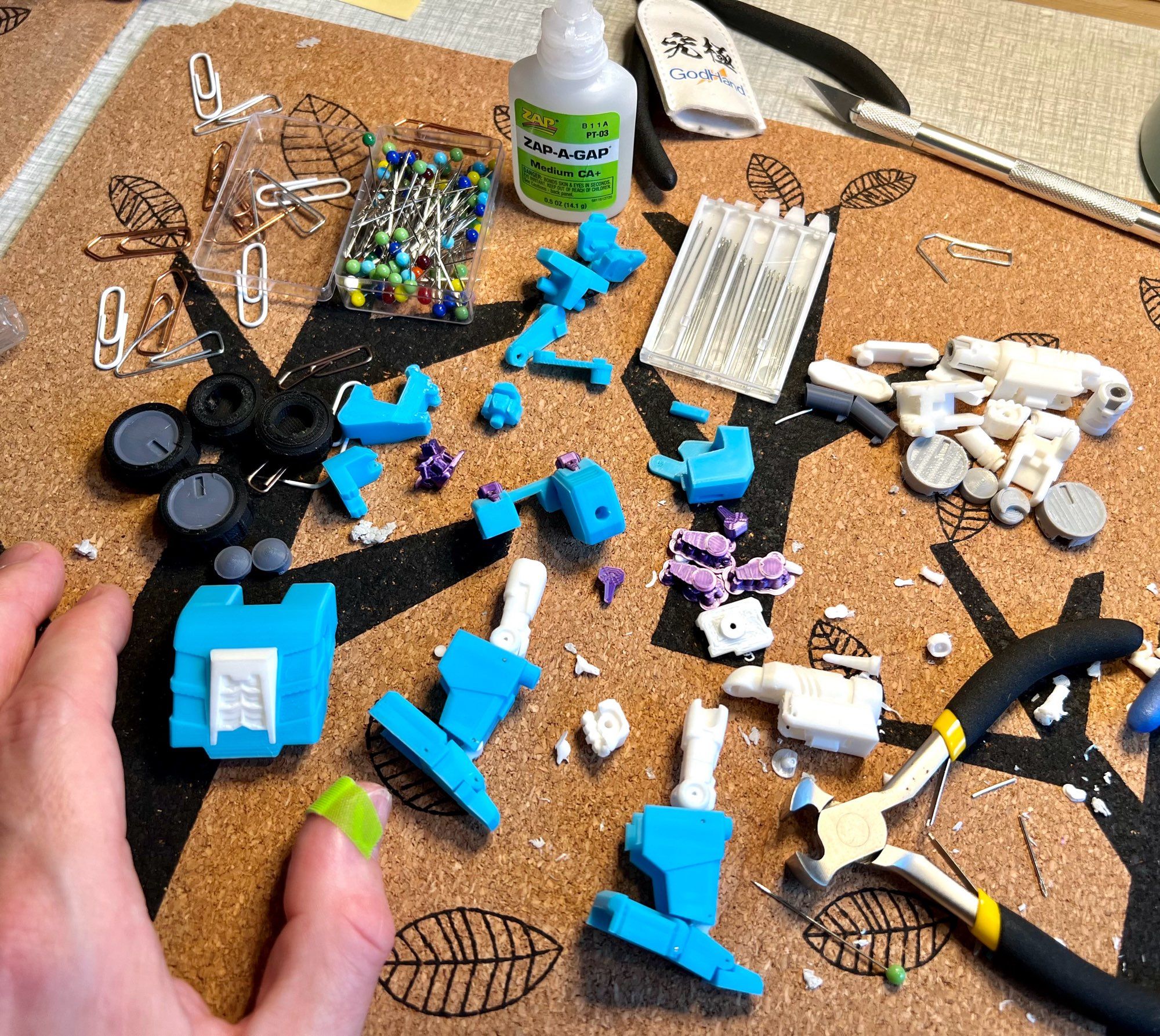 A messy desktop mat full of 3D-printed plastic parts, tools such as wire cutters and hobby knives, paper clips and needles. A hand with a band aid on the thumb rests on it